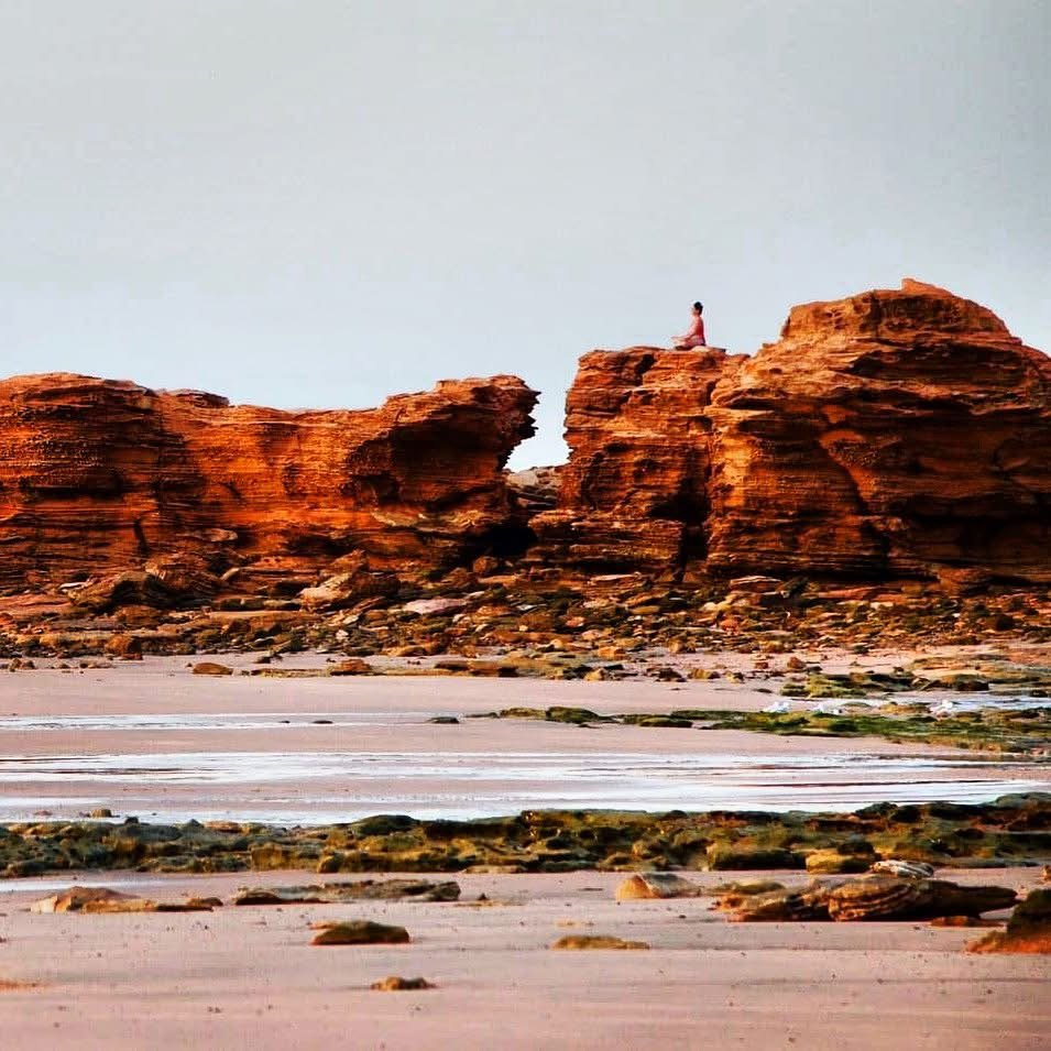 A person sitting in a yoga pose on top of large reddish-brown rock formations along a shoreline with sand, small rocks, and patches of green moss, under a clear sky.