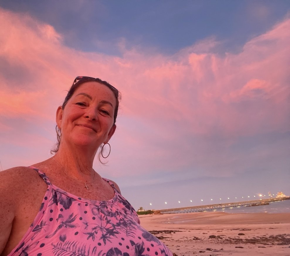 A woman smiling on a beach at sunset, with pink and purple clouds in the sky and a pier in the distance.