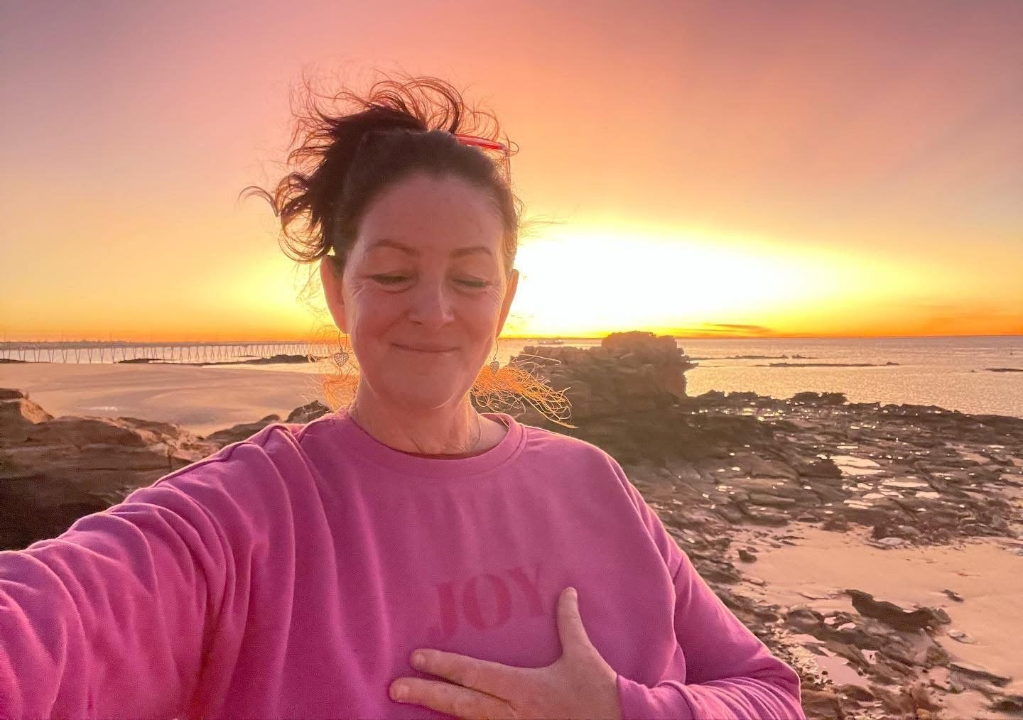 A woman taking a selfie on a rocky beach at sunset, smiling with eyes closed and hand over her chest, wearing a pink sweatshirt.