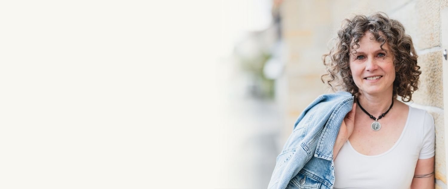 Blonder, curly-haired Frau im weißen T-Shirt mit einer Jeansjacke, die über die Schulter hängt, posiert smilend vor einer Steinmauer.