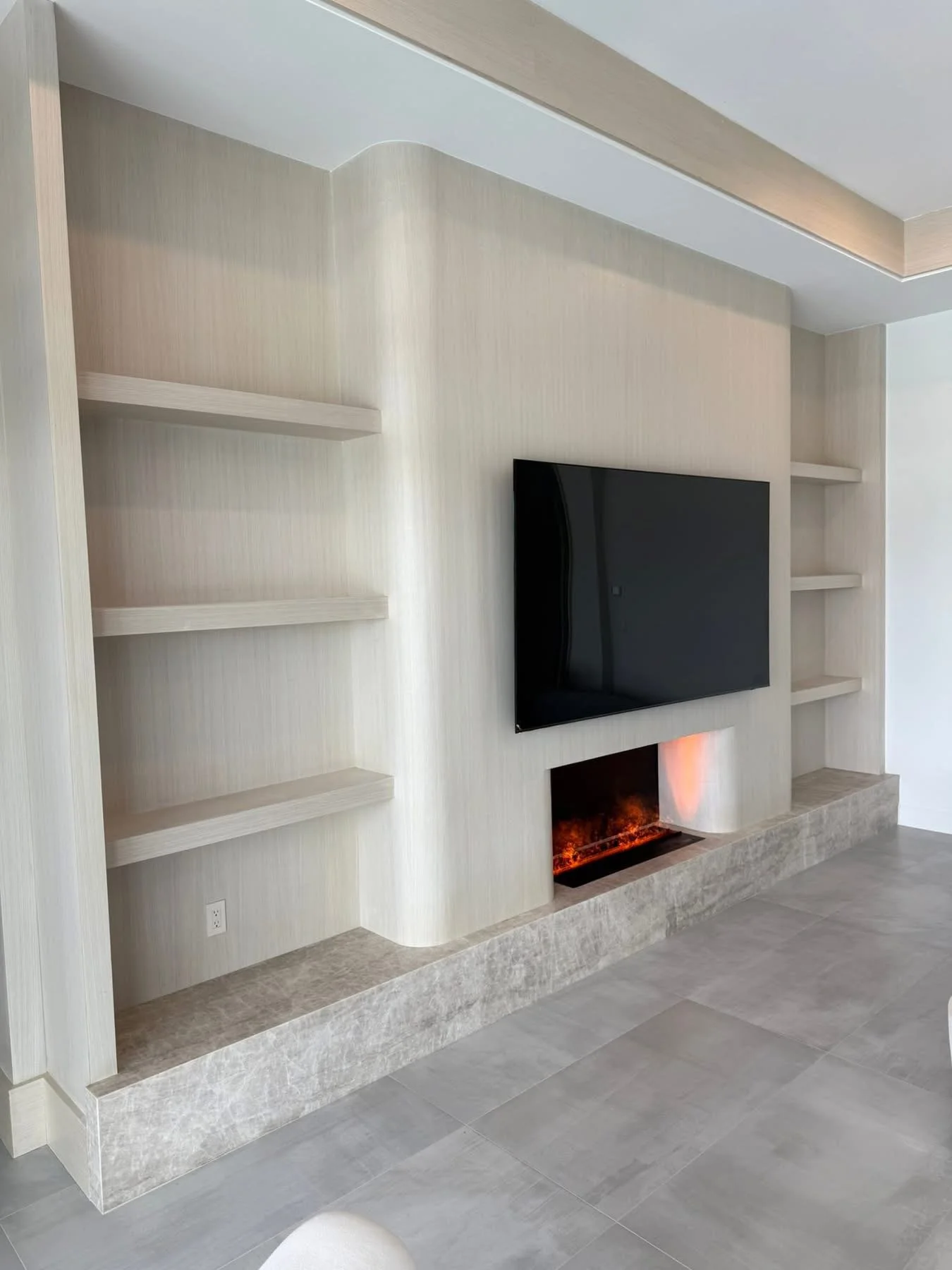 Living room with a wall-mounted flat-screen TV above a fireplace with orange flames, built-in shelves on either side, and a stone or concrete hearth at the bottom.