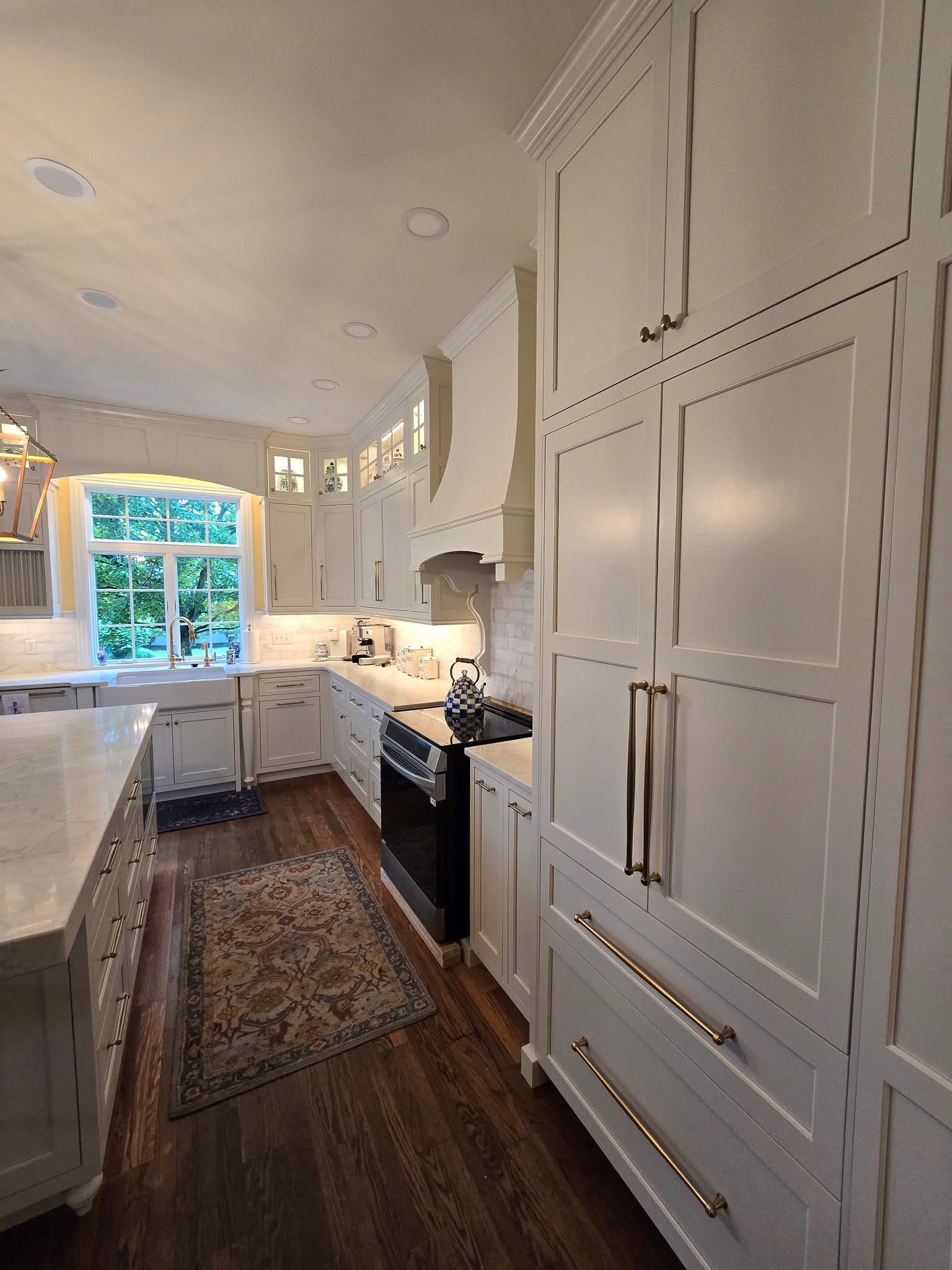 A spacious kitchen with white cabinetry, a large window above the sink, wood flooring, and a decorative rug. There are recessed ceiling lights and a white range hood over the stove. A ceramic teapot is on the stove.