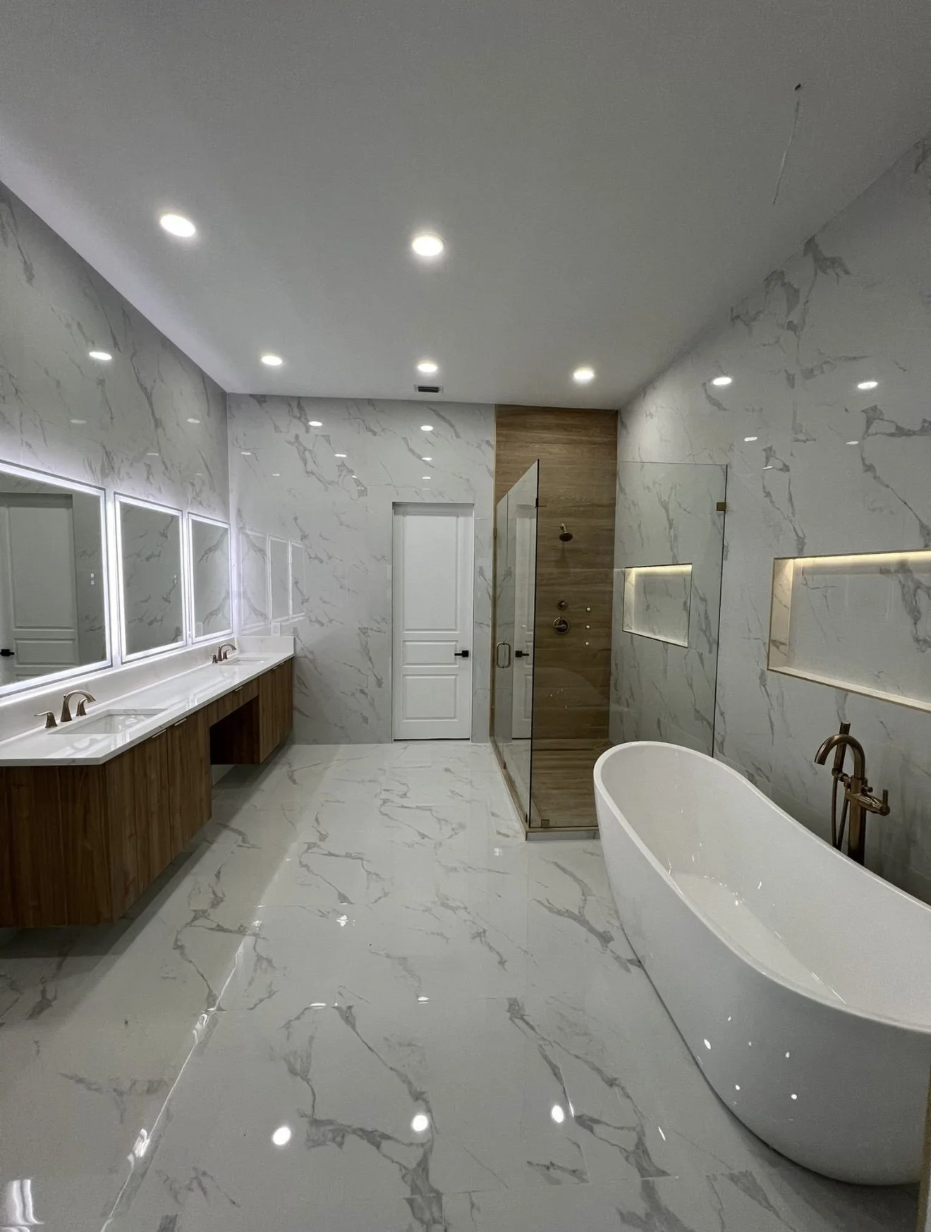 Modern bathroom with white marble floors and walls, wooden double vanity with three mirrors, white freestanding bathtub, and a shower with a glass door and wooden accent wall.