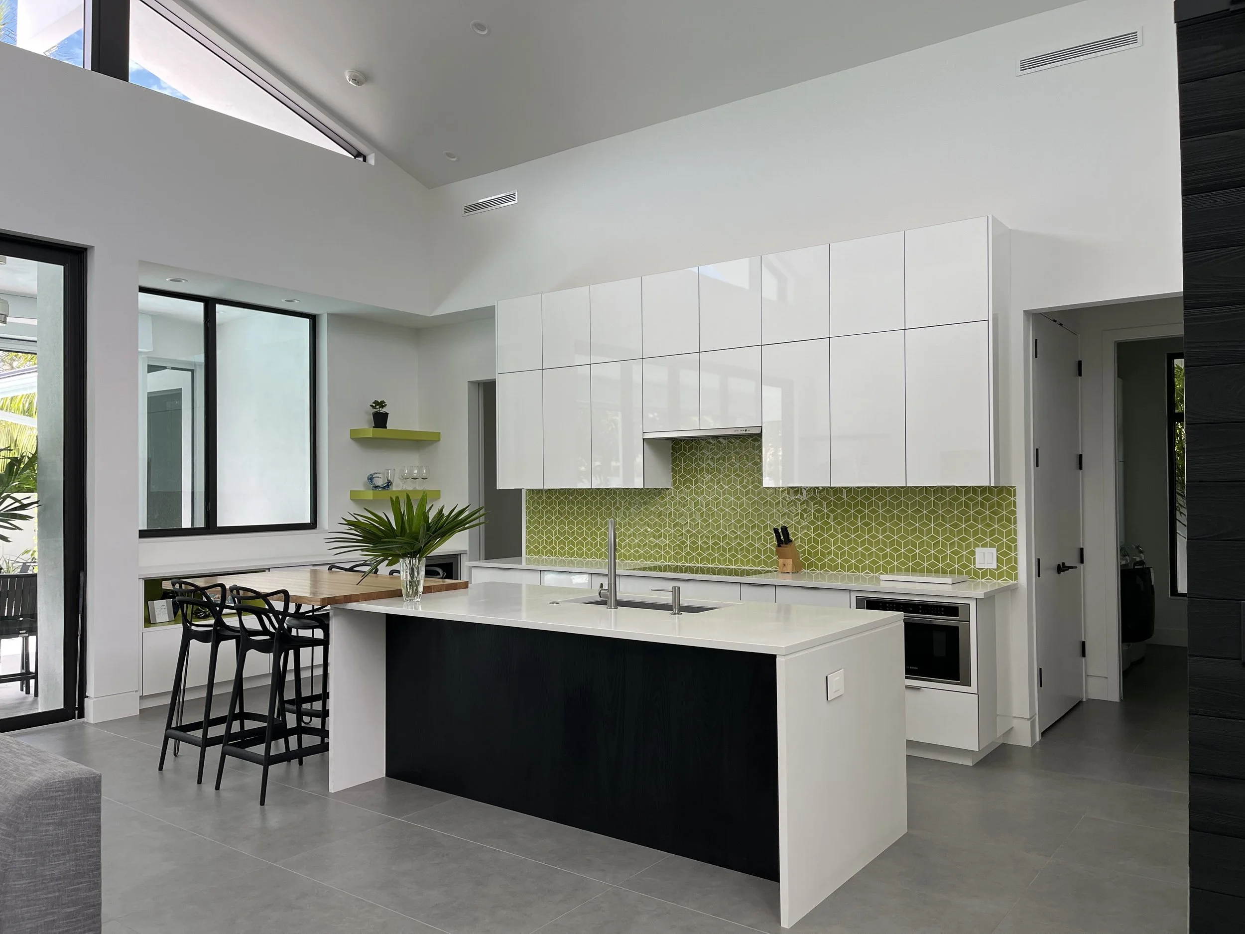 Modern kitchen with white cabinetry, green patterned backsplash, black and white island, and large windows.