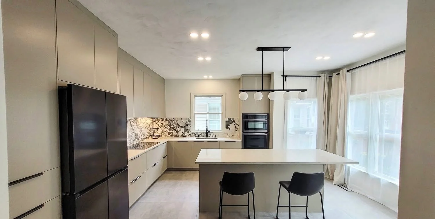 Modern kitchen with beige cabinets, black refrigerator, marble backsplash, small window above the sink, central island with white countertop, black chairs, large windows with sheer curtains, ceiling lights, and a black pendant light fixture.