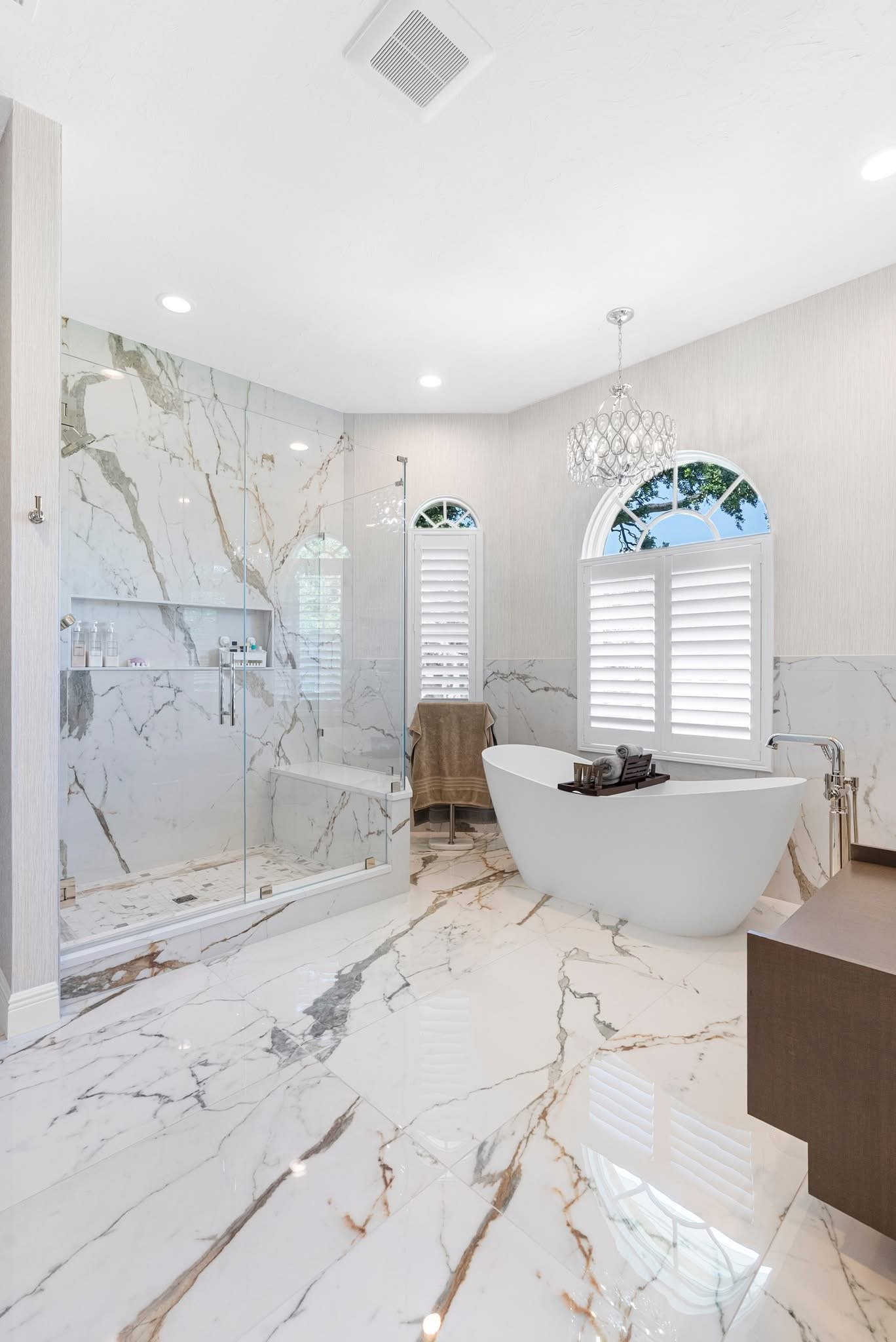 A luxurious bathroom with marble floors and walls, a glass-enclosed shower, a freestanding bathtub, and large arched windows with white shutters. There is a chandelier hanging from the ceiling.