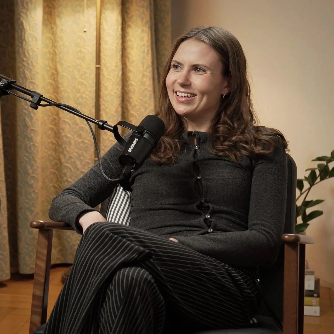 Frau sitzt entspannt in einem Gespräch, spricht in ein Podcast Mikrofon, lächelt und trägt einen schwarzen Pullover mit Knöpfen, im Hintergrund goldene Vorhänge und Pflanzen.