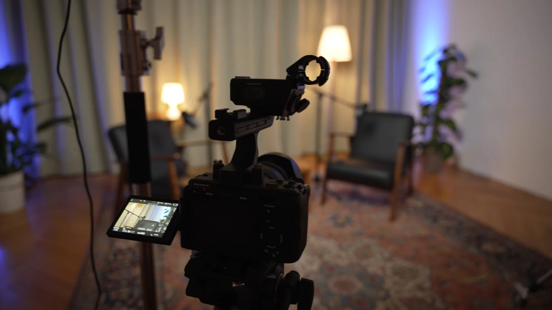 Eine Kamera auf einem Stativ in einem Podcast Studio mit einem Sessel, einer Stehlampe und Pflanzen, vorbereitet für Videoaufnahmen.