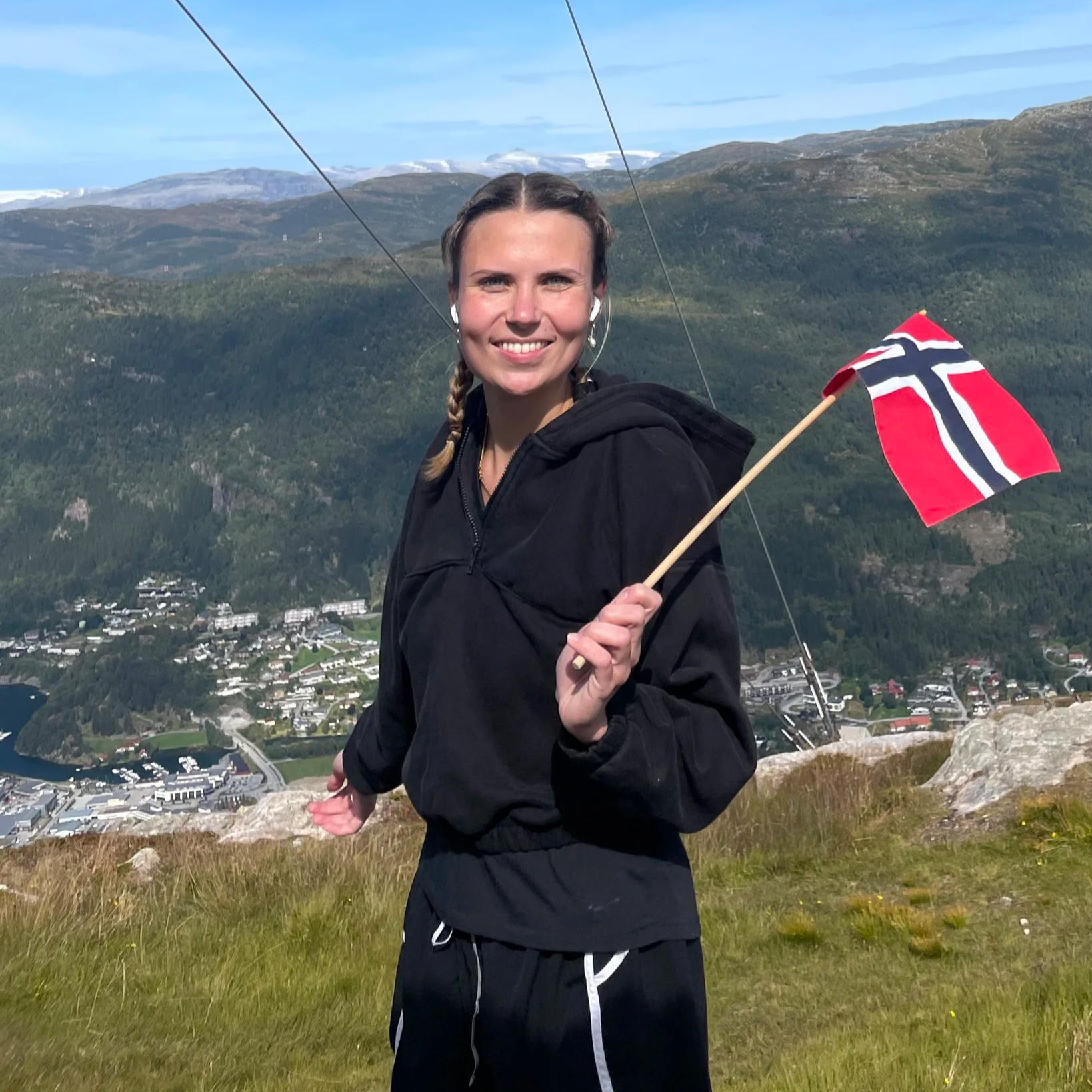 Eine lachende Frau mit Zopf hält eine norwegische Flagge auf einem Hügel mit Blick auf eine Stadt und Berge im Hintergrund.