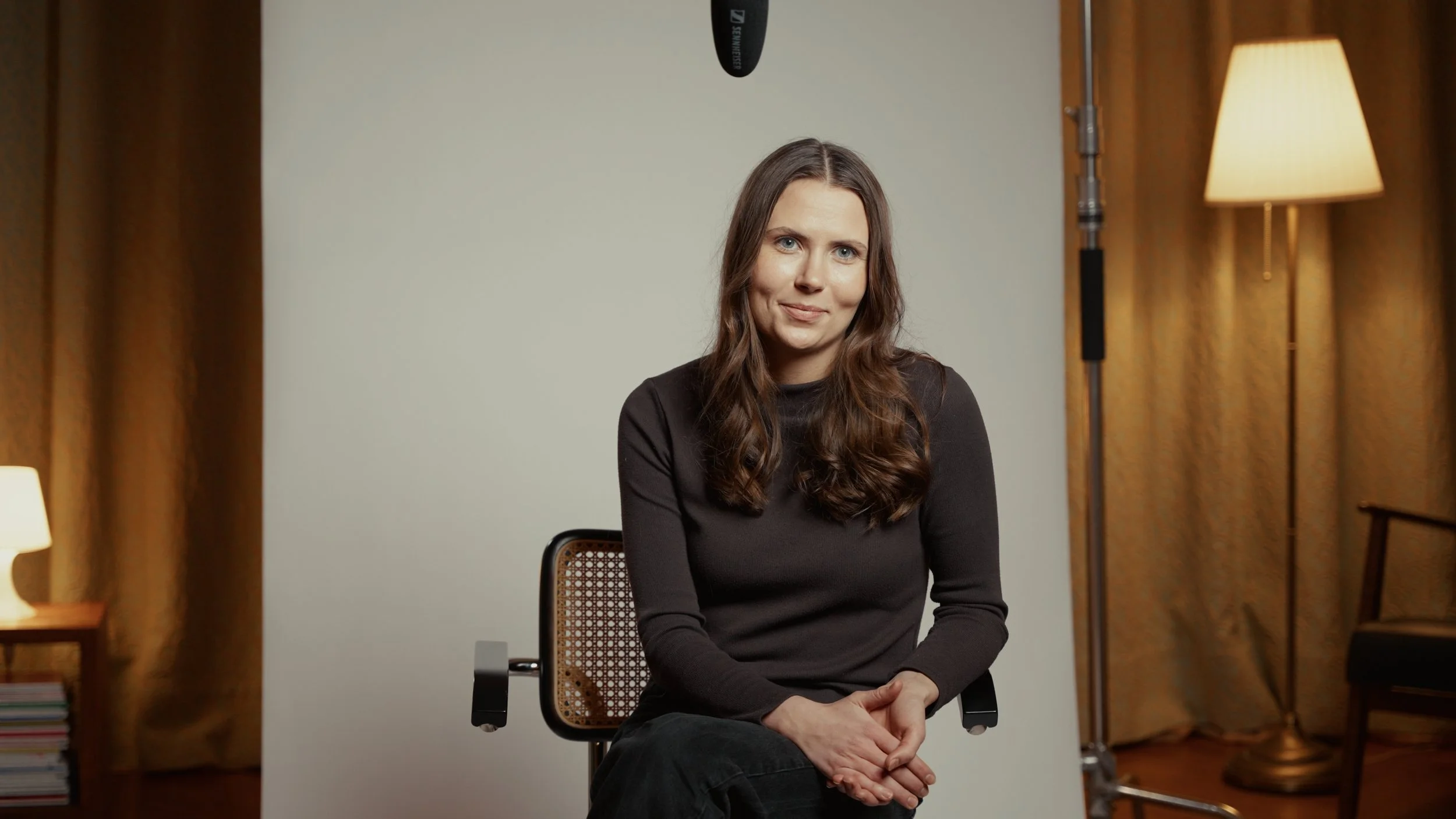 Frau sitzt vor einem weißen Hintergrund in einem Interview-Setup mit Lichtquellen und einer Kurzhintergrundkulisse, lächelt leicht.