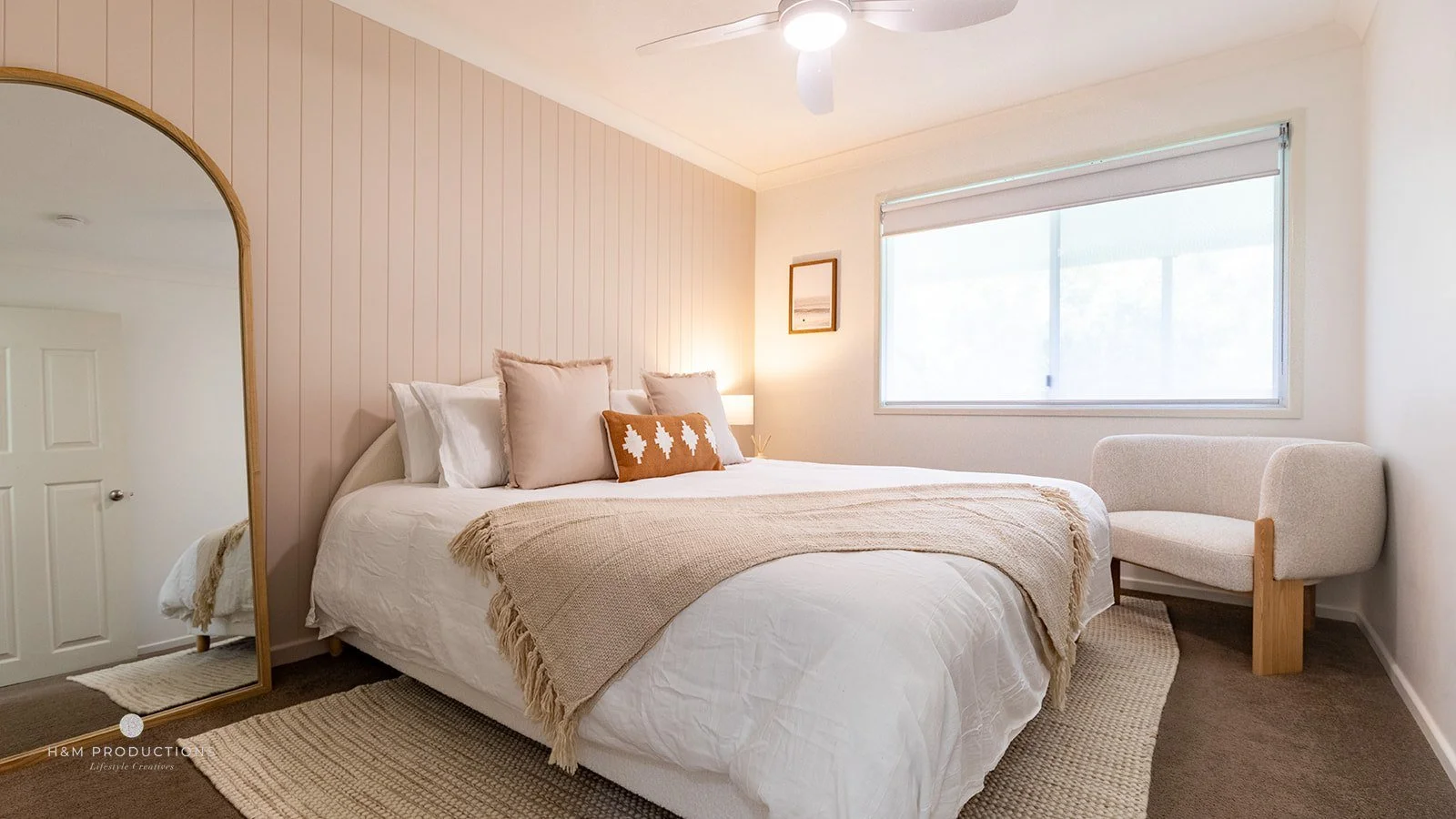 A neatly made bed with beige and white pillows, a tan throw blanket, and a window letting in natural light, in a minimalistic bedroom with light-colored walls and a framed picture hanging.