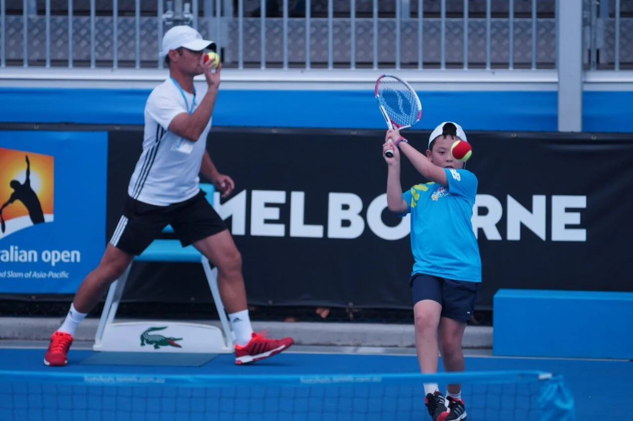 A young boy practicing tennis on an outdoor court with an adult. The boy is holding a tennis racket and ball, wearing a white cap, blue shirt, and black shorts. The adult behind him is also holding a tennis ball, dressed in a white shirt, black shorts, and red shoes. There is a sign in the background that says "Melbourne" and a banner with