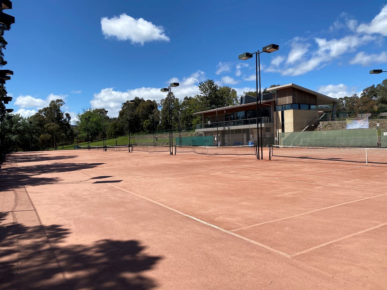 Empty outdoor tennis courts with court lines, net, surrounding fencing, and a nearby building, under a partly cloudy blue sky with trees in the background.