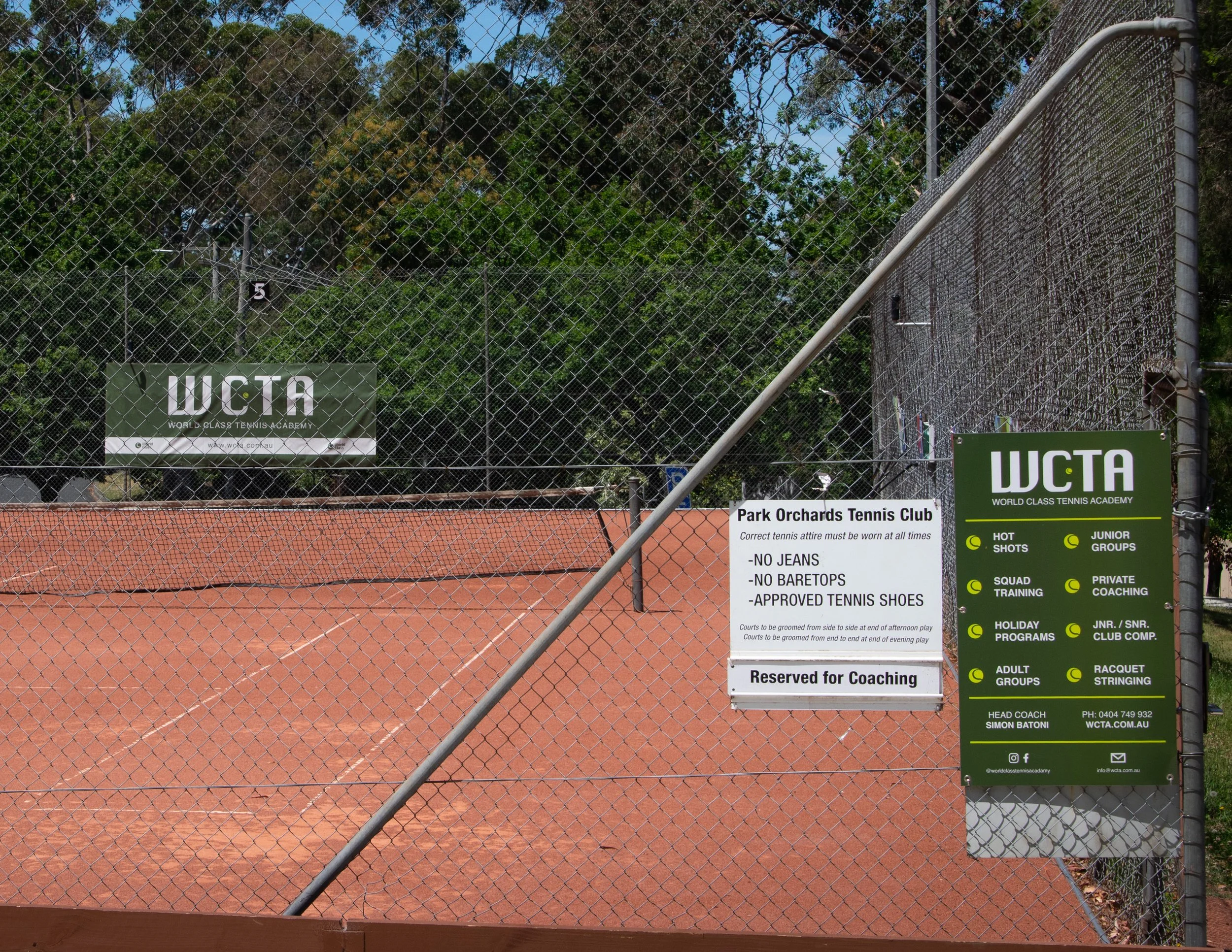 Fenced tennis court with signs for World Class Tennis Academy, including instructions and rules for dress code, coaching reservation, and club information, surrounded by trees and a blue sky.