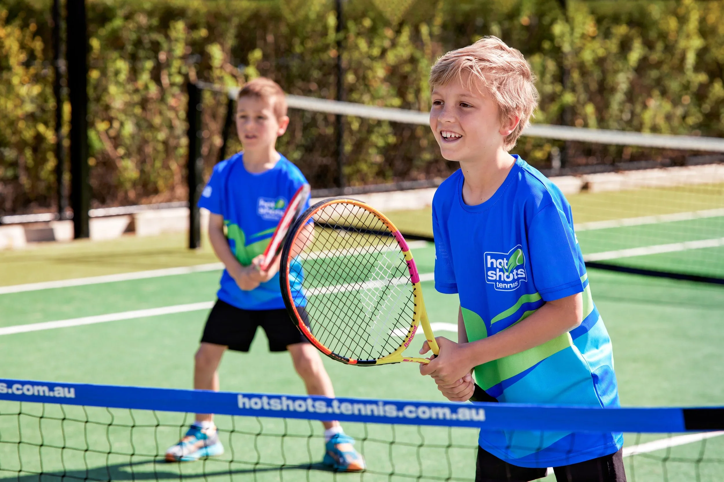 Children playing tennis on an outdoor court, with one girl in the foreground holding a tennis racket and a tennis ball on the ground, other children in the background, and green trees behind a chain-link fence.
