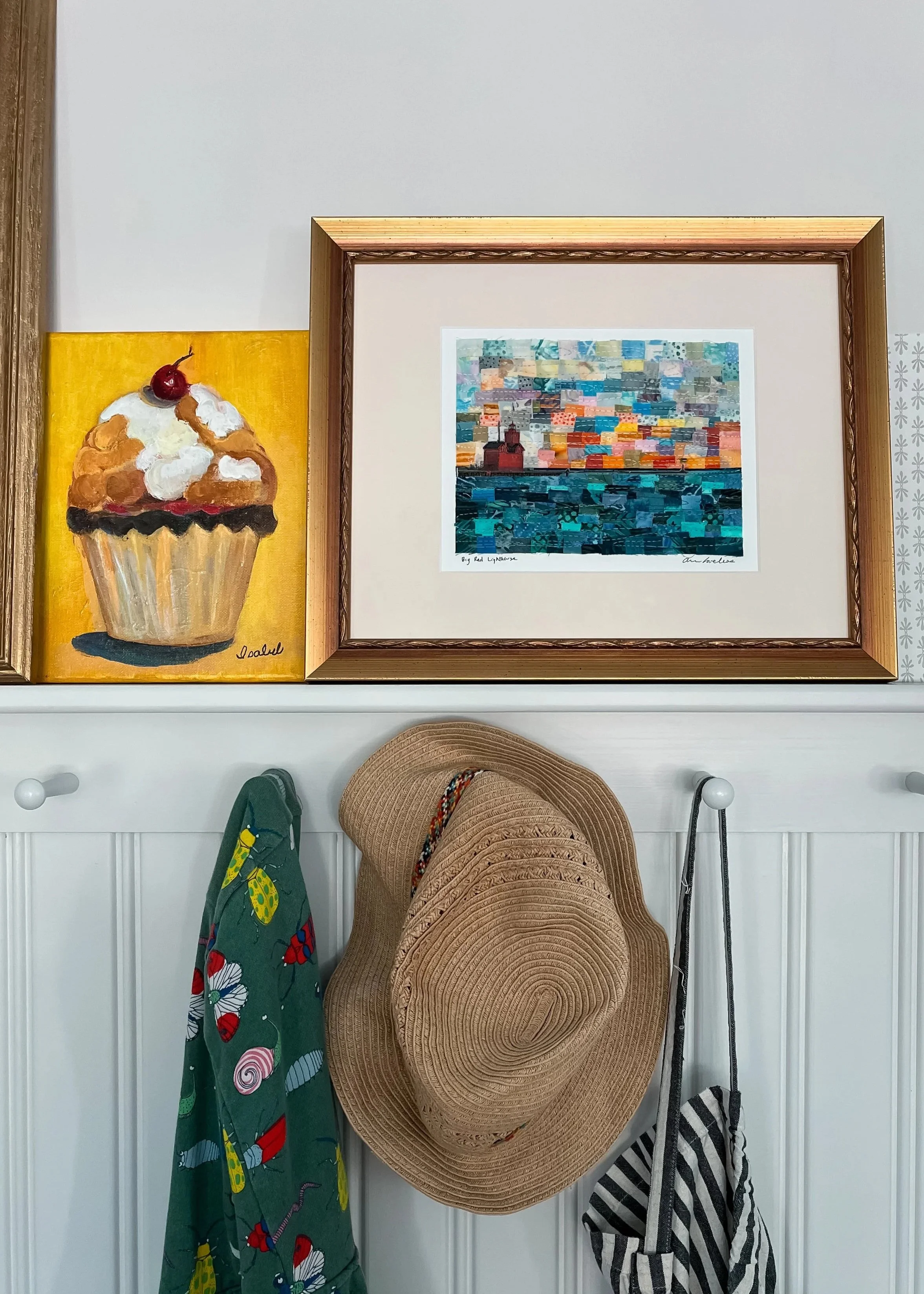 Paintings hanging on a wall above a white wainscoting. One is a portrait of a cupcake with a cherry on top, on a yellow background. The other is a framed abstract, colorful seascape, with a wooden frame. Below, hats and bags are hanging on white hooks.