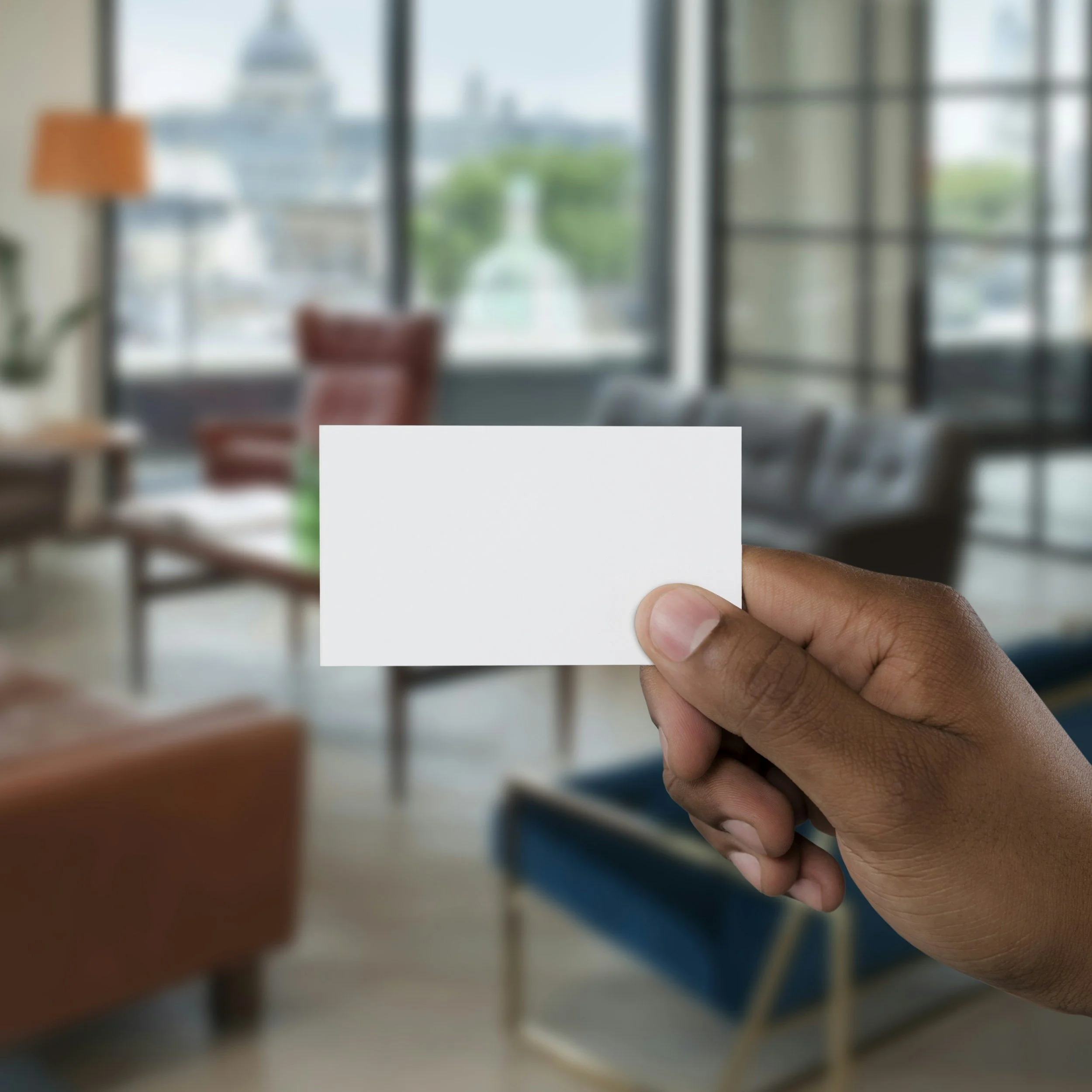 A hand holding a blank white business card against the backdrop of a room representing the decision therapists need to make about using business cards for their private practice