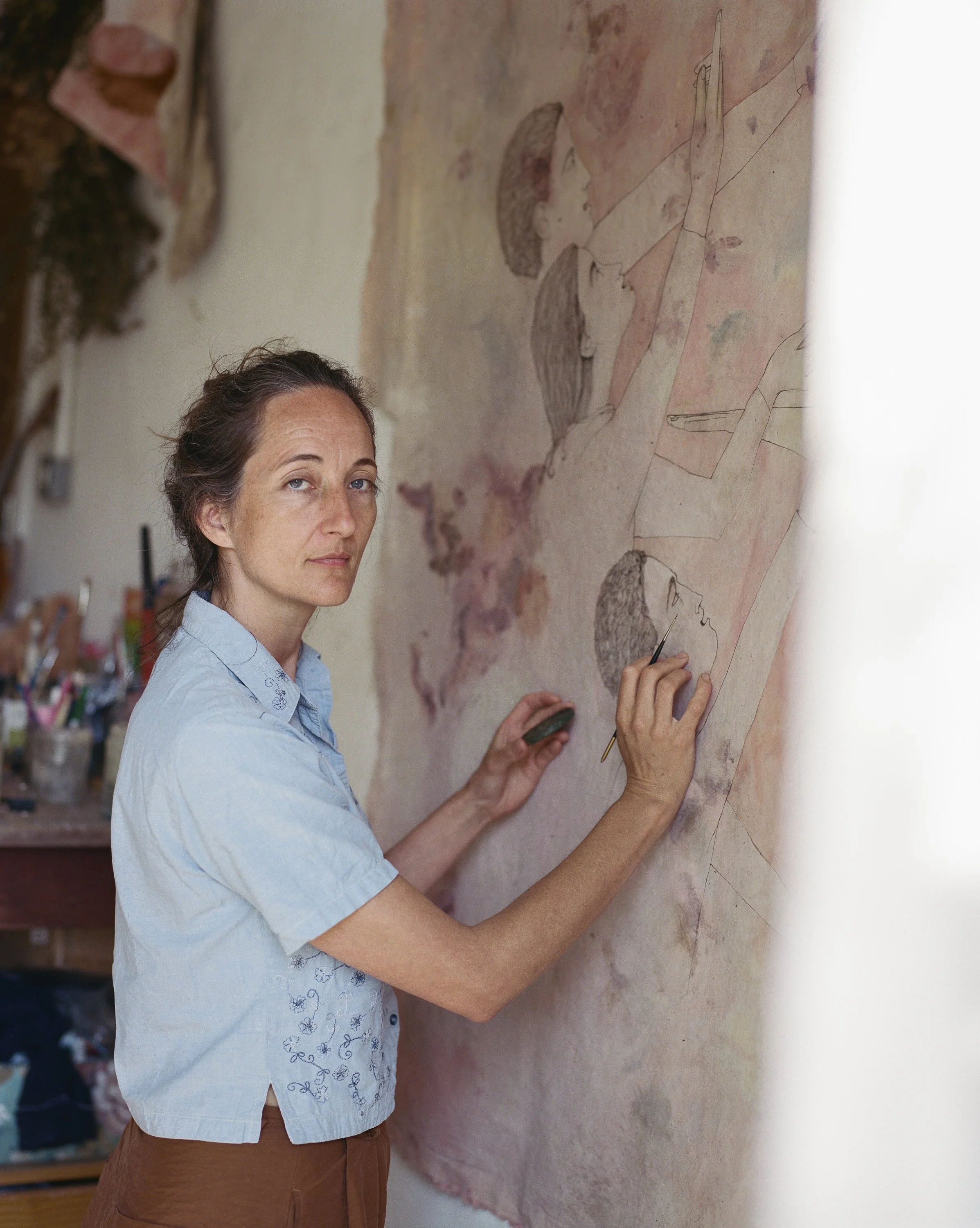 A woman in a light blue shirt working on a mural or wall painting, which features sketches of hands and faces in a classical or mythological style.