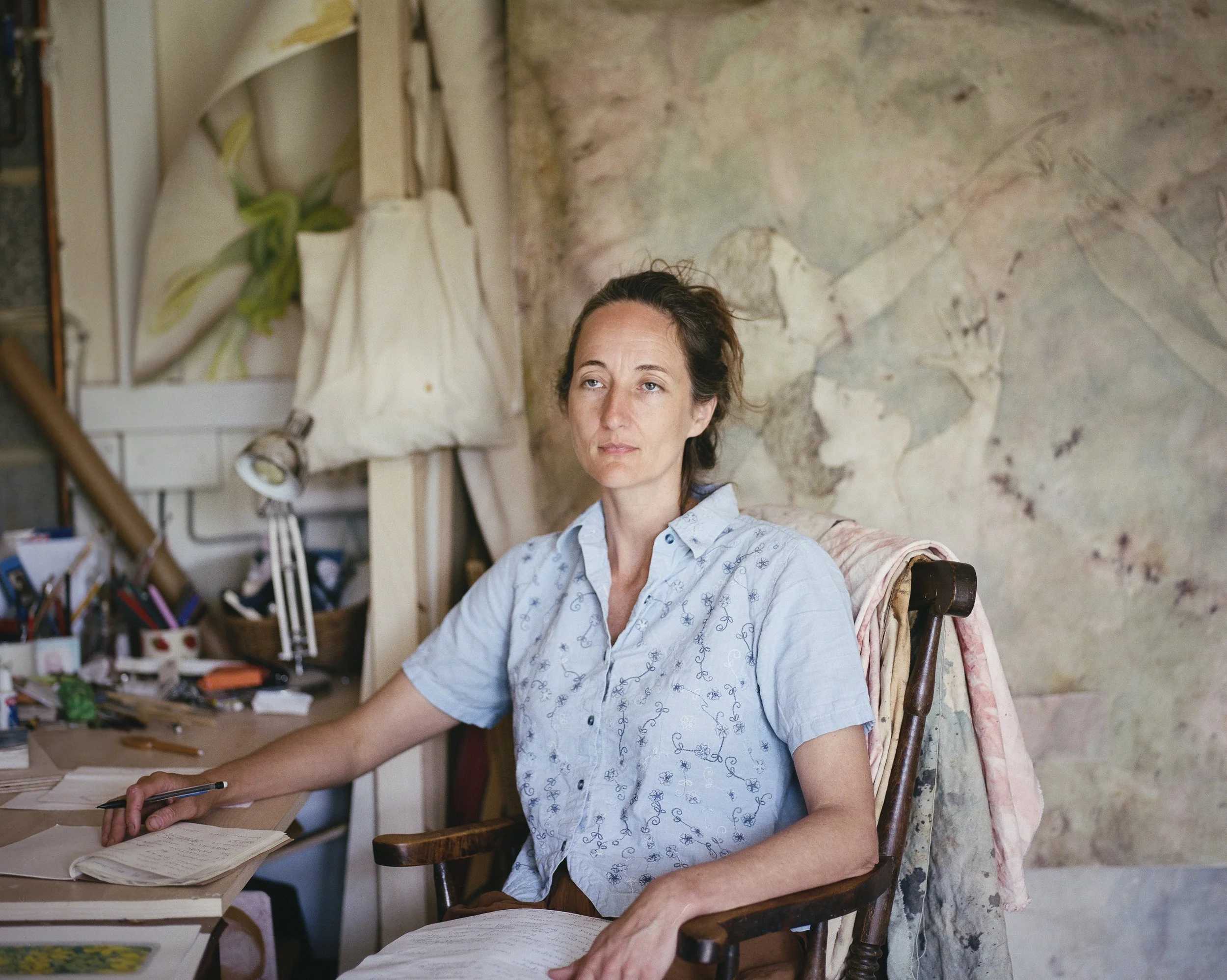 A woman sits at a cluttered desk in a room with a textured, abstract mural on the wall behind her. She is wearing a light blue embroidered shirt and has her hair pulled back.