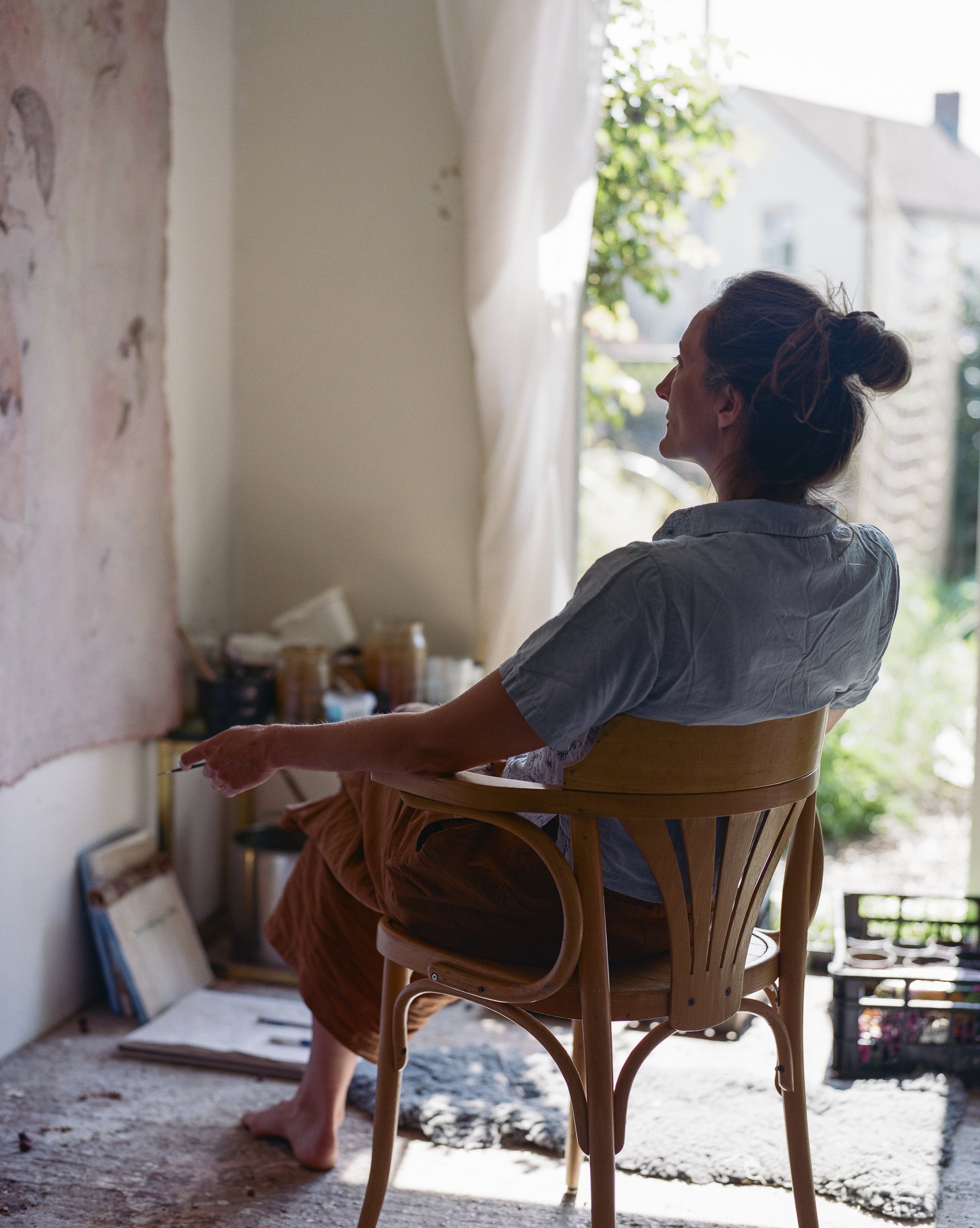 A woman sitting on a wooden chair near a window, looking outside with a relaxed posture.