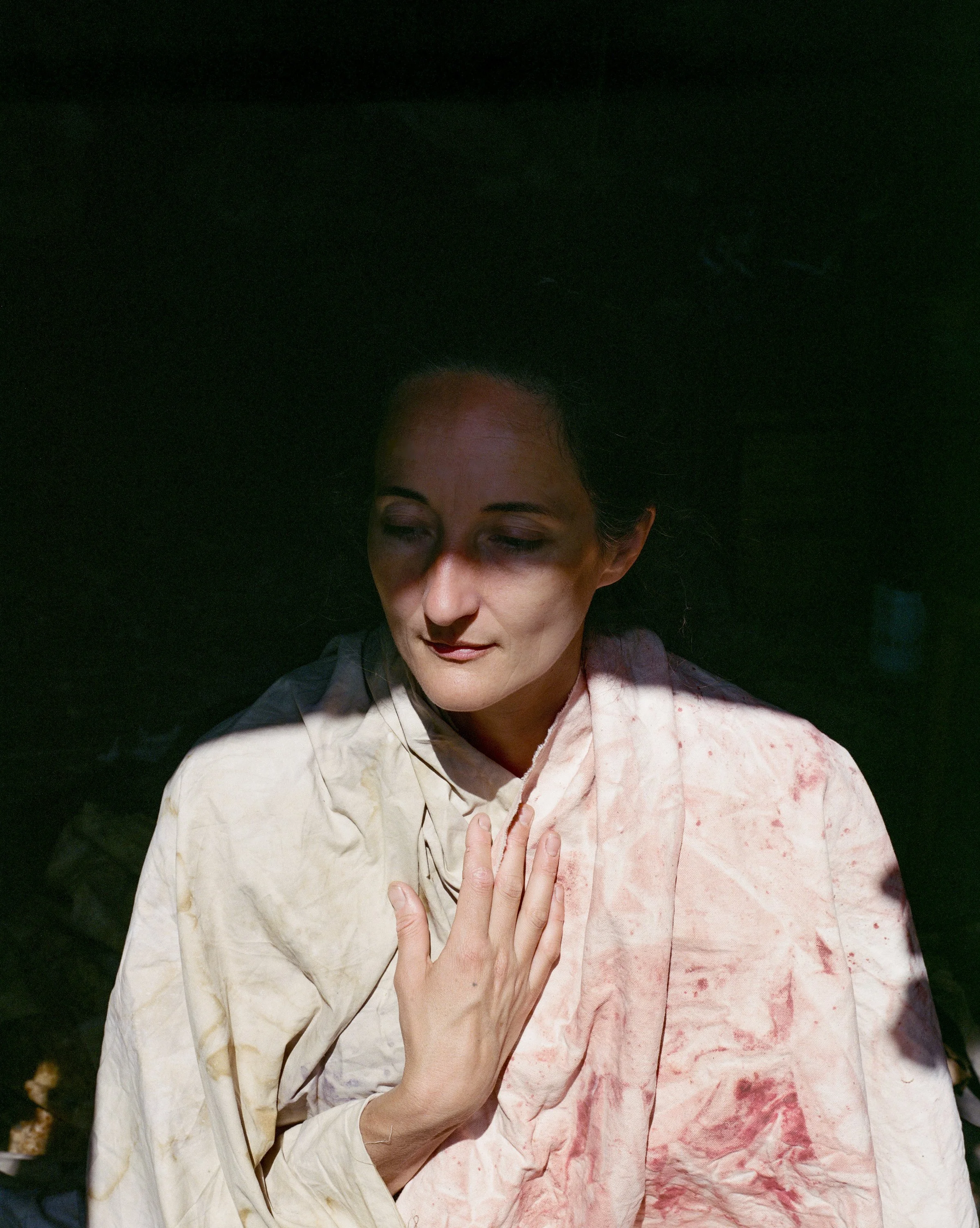 A woman with dark hair and pale makeup, gazing downward, dressed in a light-colored, stained cloth with her hand on her chest, against a black background.