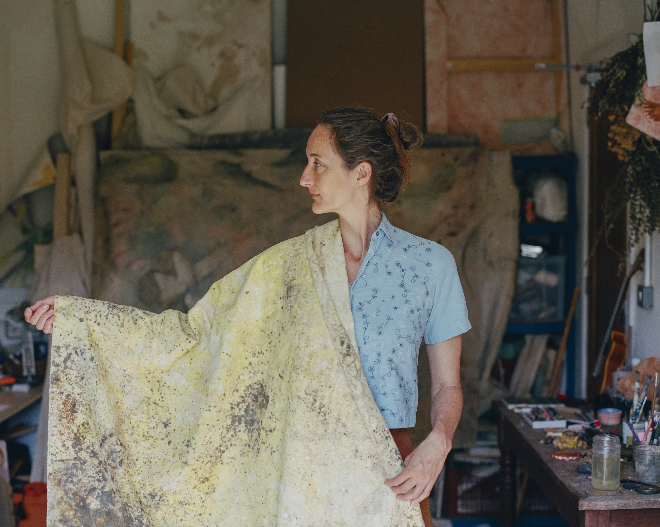 A woman in a short-sleeved blue shirt stands in an art studio holding a large piece of abstract artwork. The studio has a cluttered workspace with various art supplies, jars, and brushes on a wooden table, and a colorful canvas or backdrop in the background.