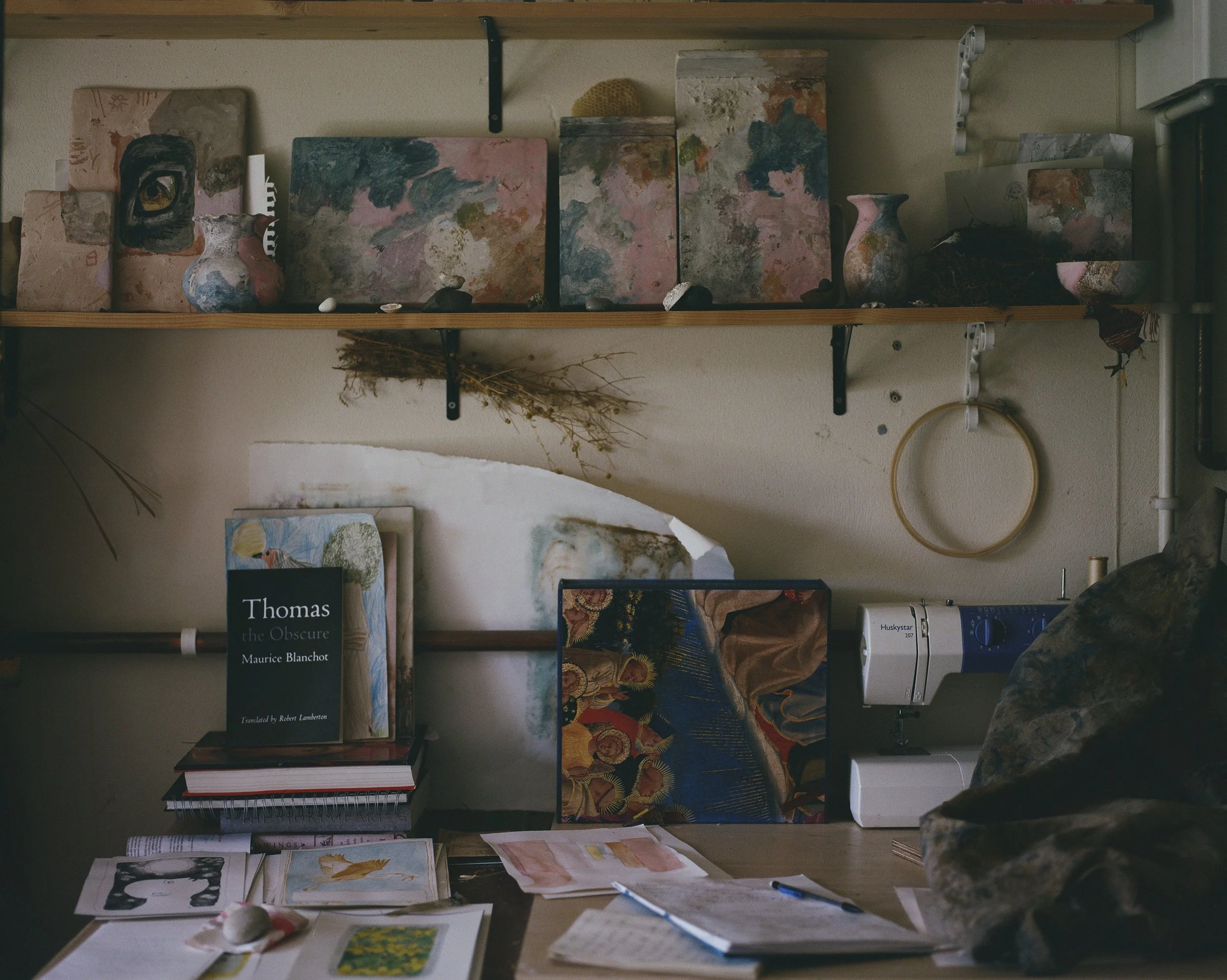 An art studio workspace with shelves holding abstract paintings and vases, a table with books including Maurice Blanchot's 'Thomas the Obscure', sketches, and art supplies, and a sewing machine nearby.