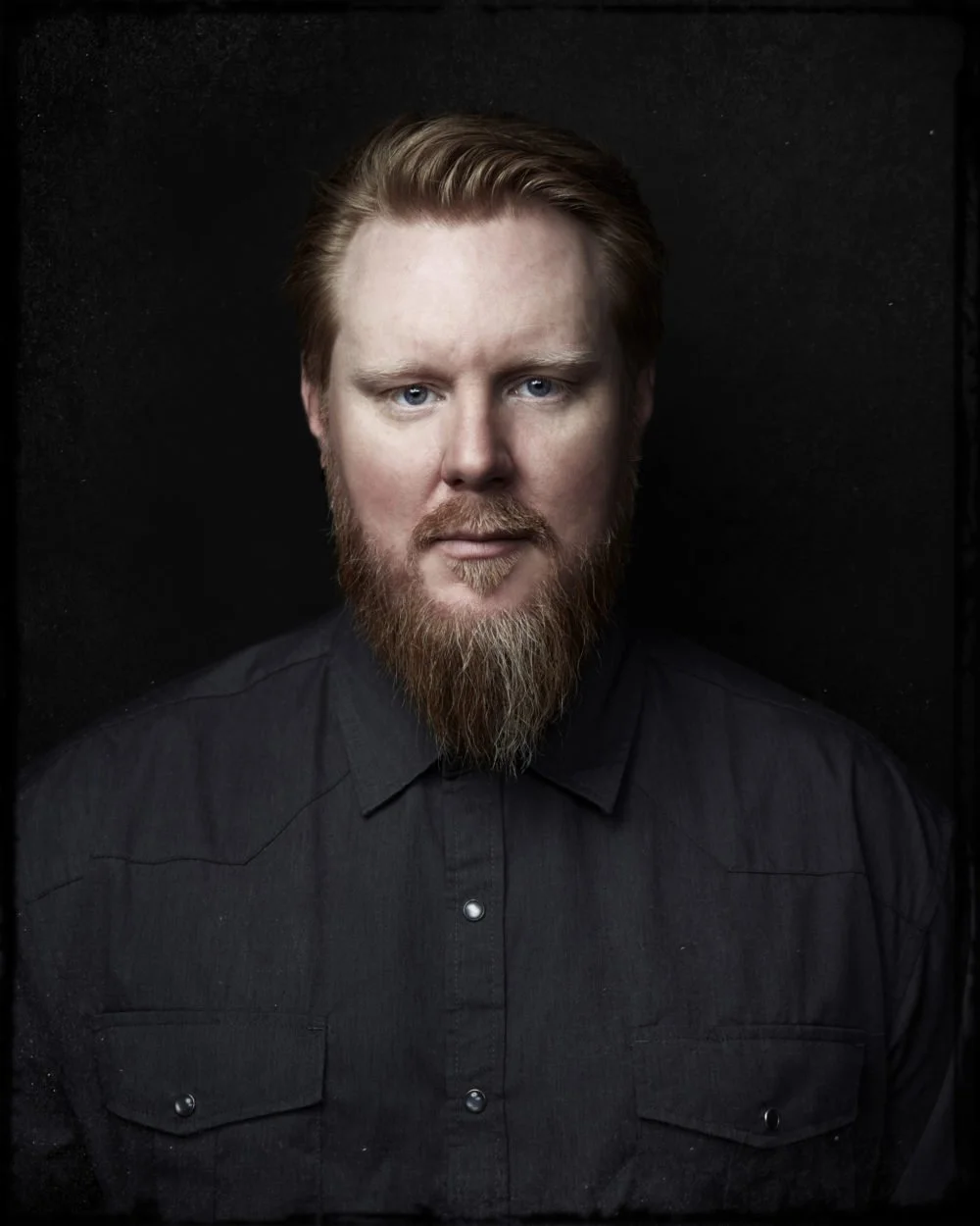 Portrait of a man with red hair and beard wearing a black shirt, set against a black background.