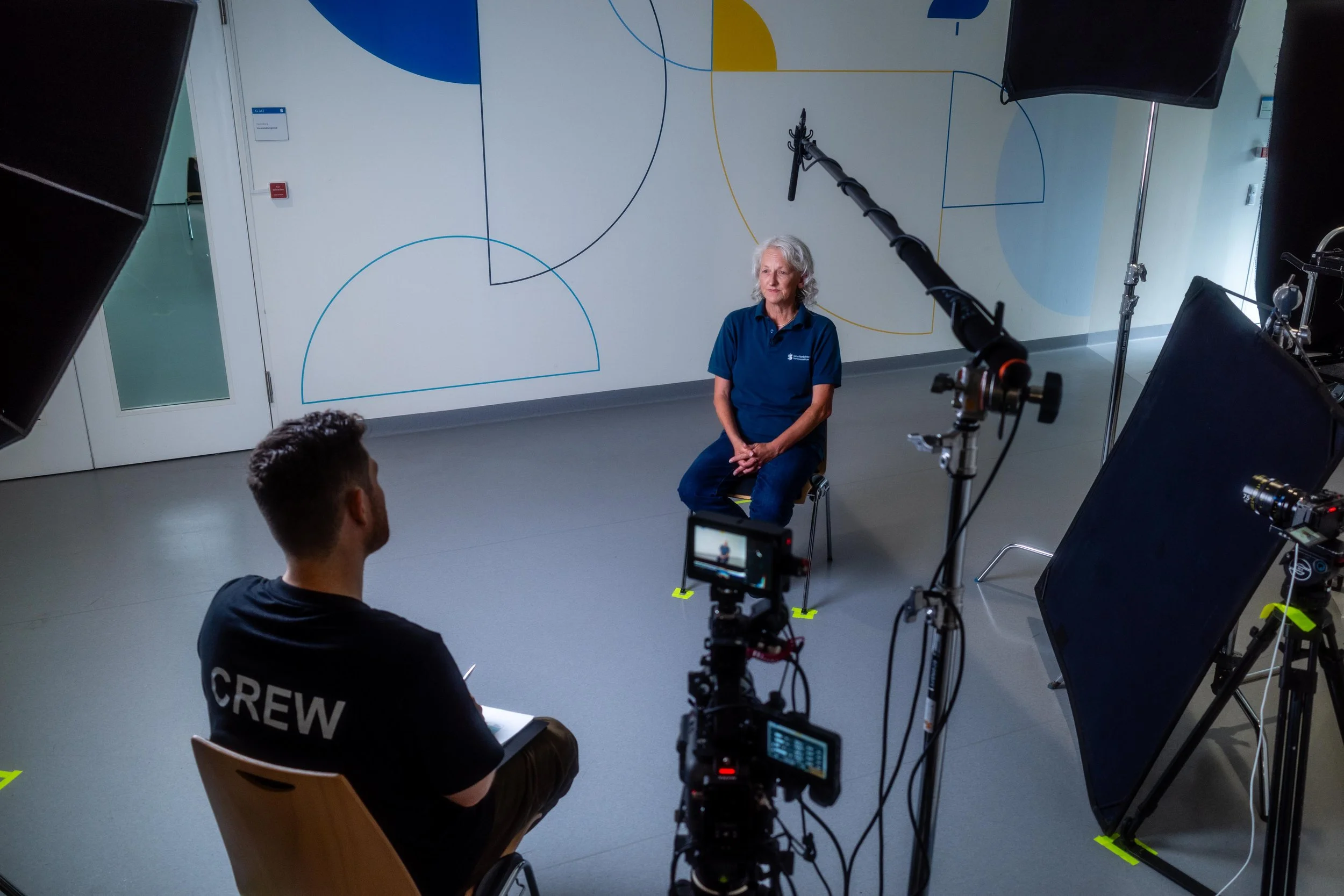 Eine Frau sitzt in einem Studio vor einer weißen Wand mit bunten Linien, während ein Filmteam sie filmt. Das Team besteht aus einer Person mit 'CREW'-Aufnäher auf dem Rücken, die eine Kamera bedient, und weiteren technischen Ausrüstungen, darunter Beleuchtung und Mikrofone.