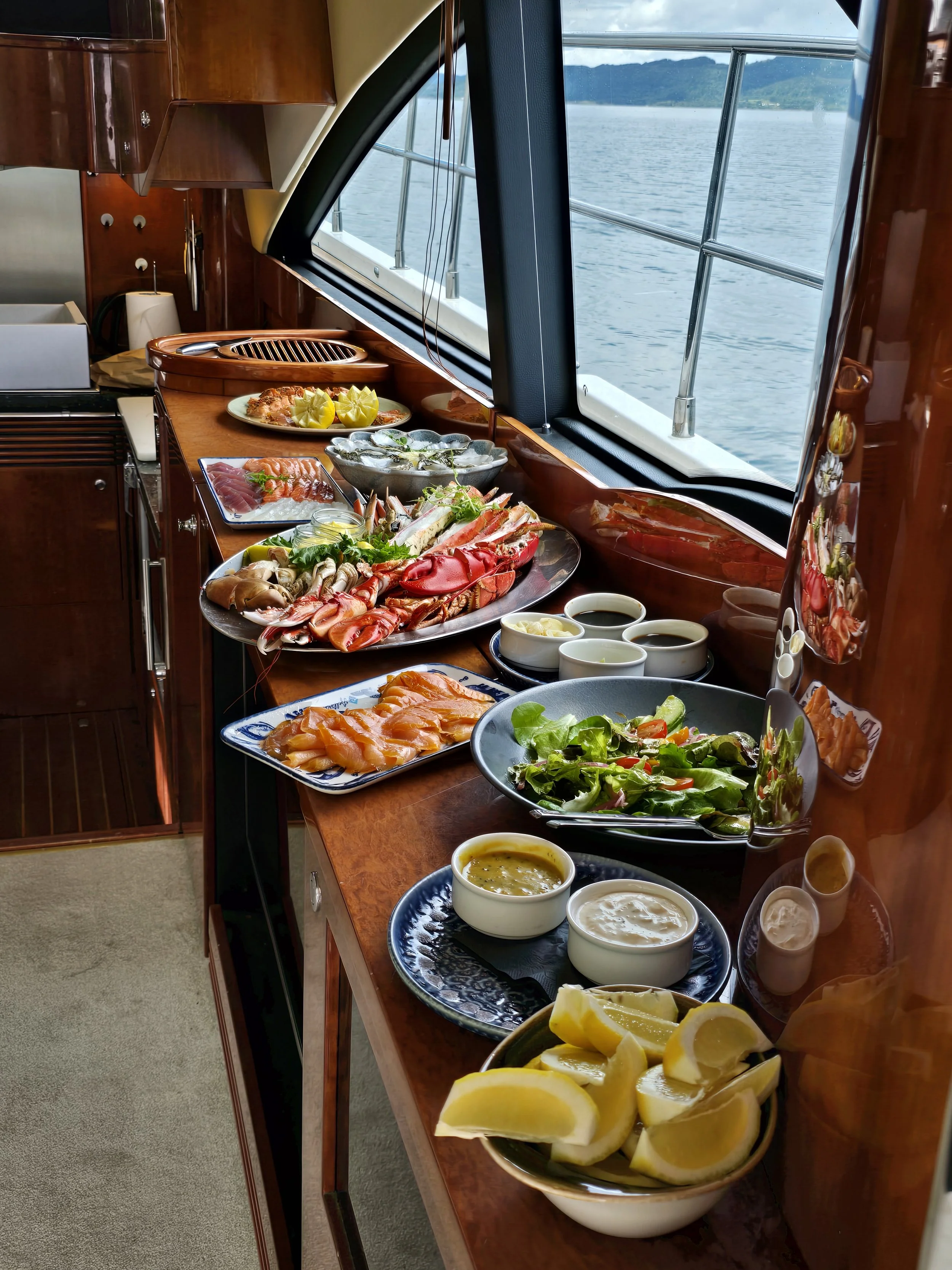 Assorted seafood, salads, lemon slices, and dipping sauces arranged on a dining table on a boat near a window with a view of the water.