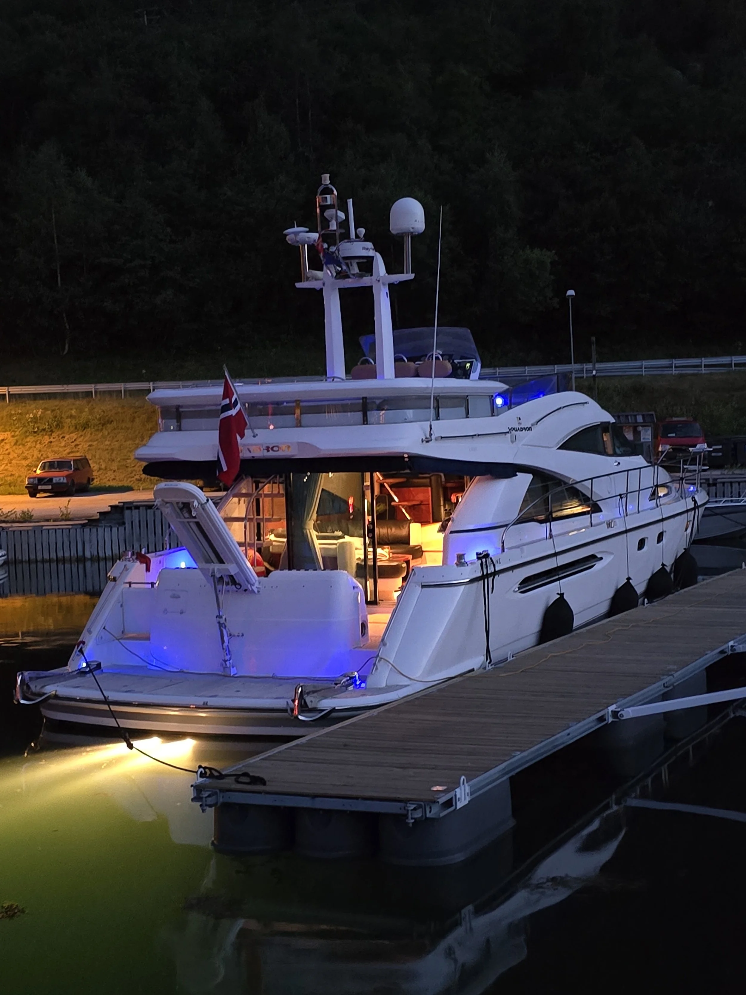 A white yacht docked at a marina during evening with blue and yellow lighting, Canadian flag flying, and a background of trees and parked cars.