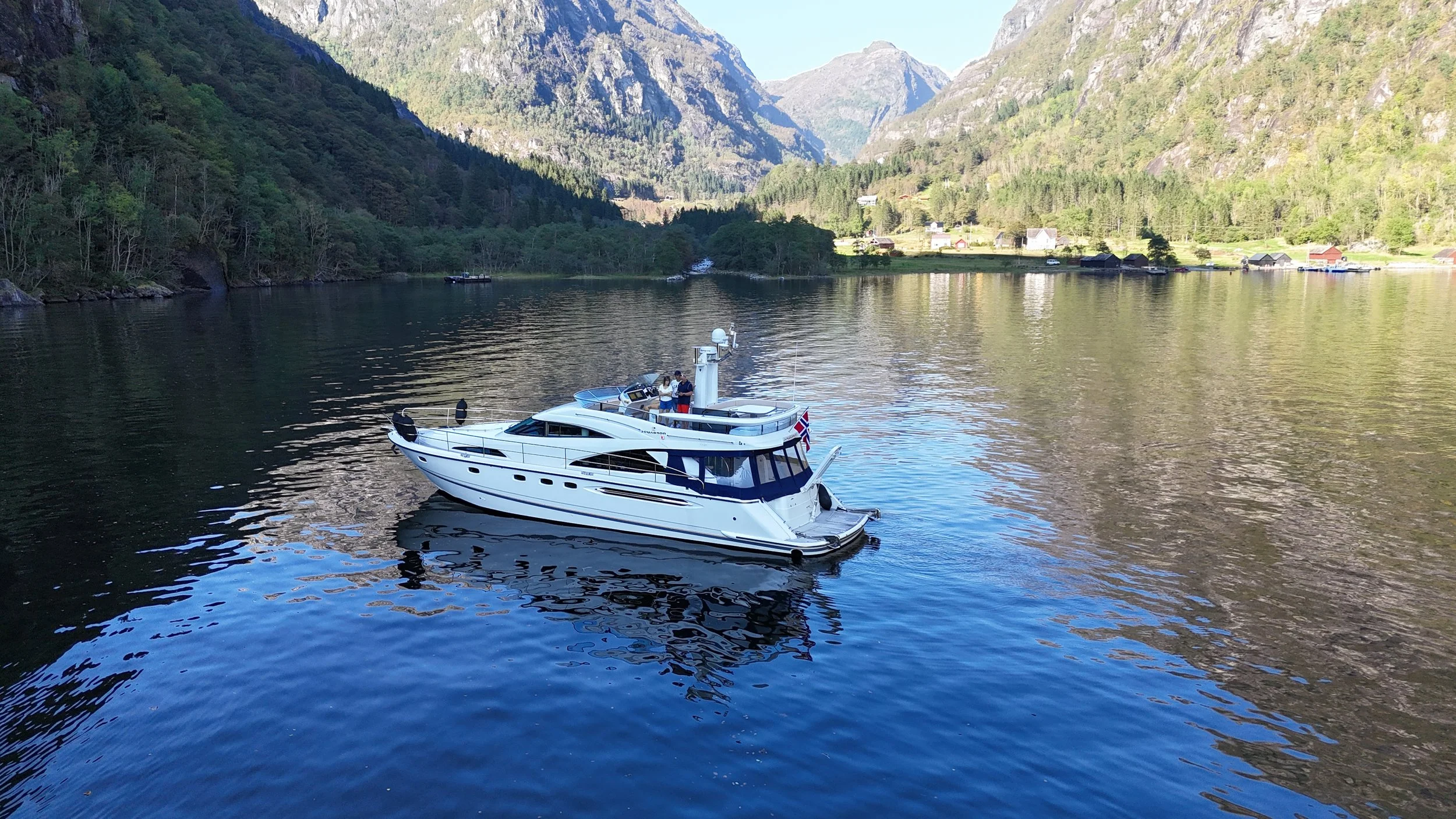 A white yacht sailing on a calm, reflective body of water surrounded by green mountains and a small village in the background.