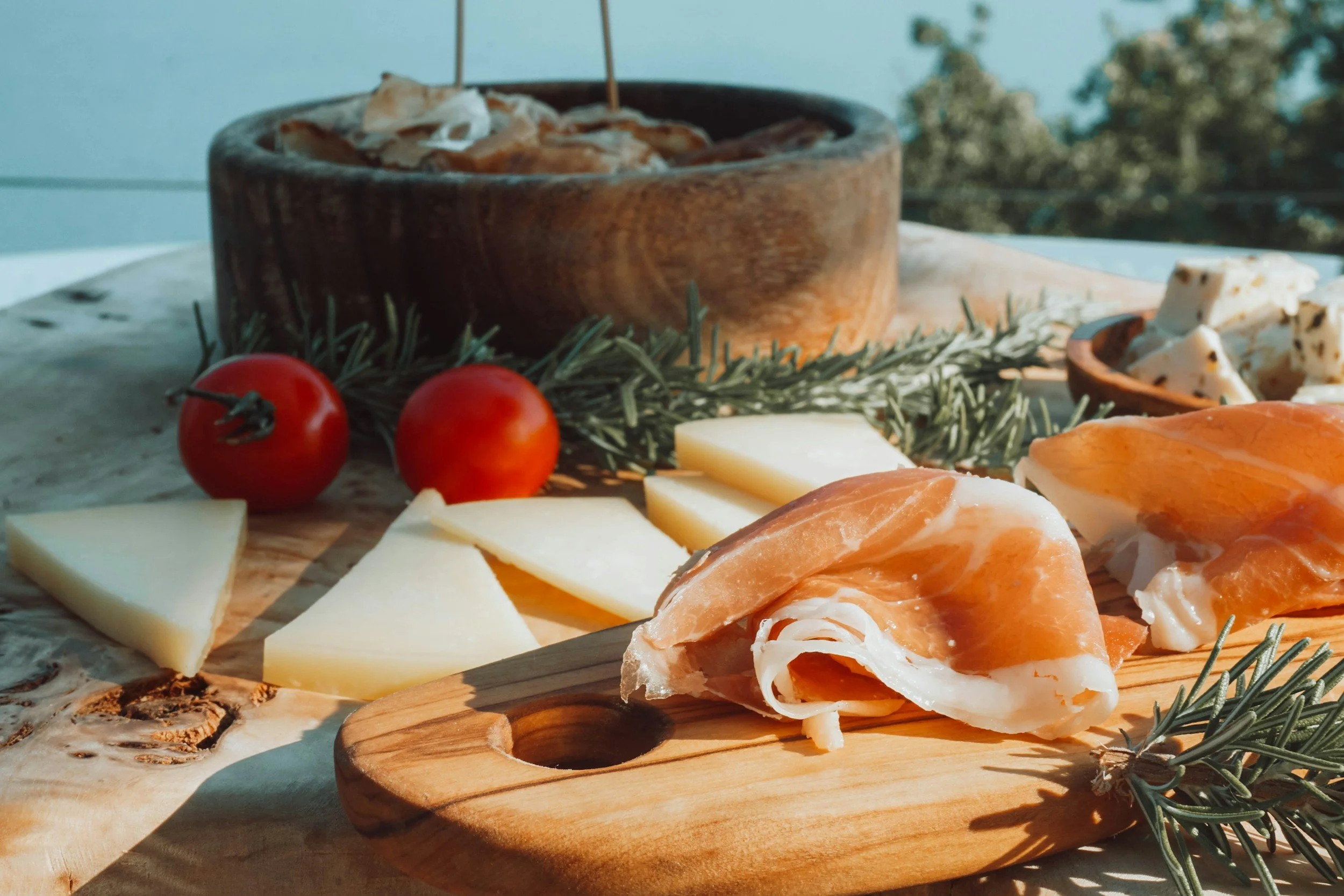Assorted cheeses including slices and wedges on a wooden cutting board, fresh cherry tomatoes, sprigs of rosemary, and a charcuterie board with sliced cured meat on a rustic outdoor table.
