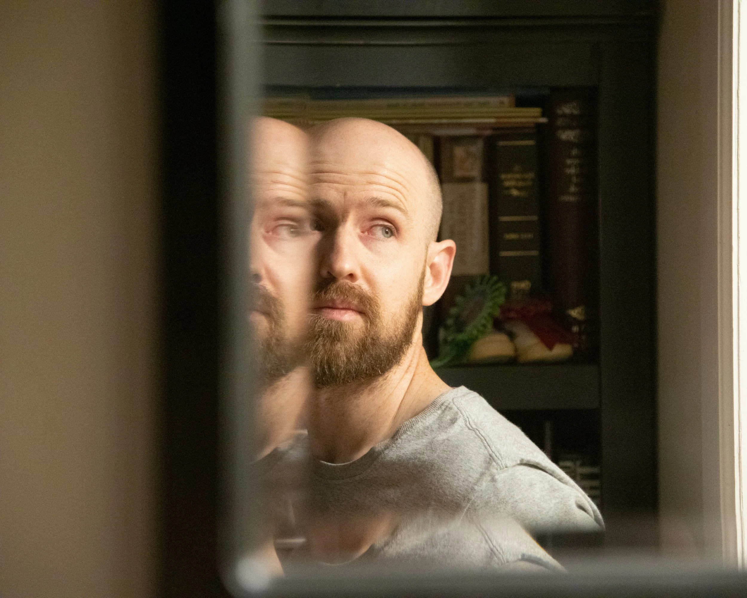 Close-up of a man's face seen through a gap in a door or wall, with a bookshelf in the background.