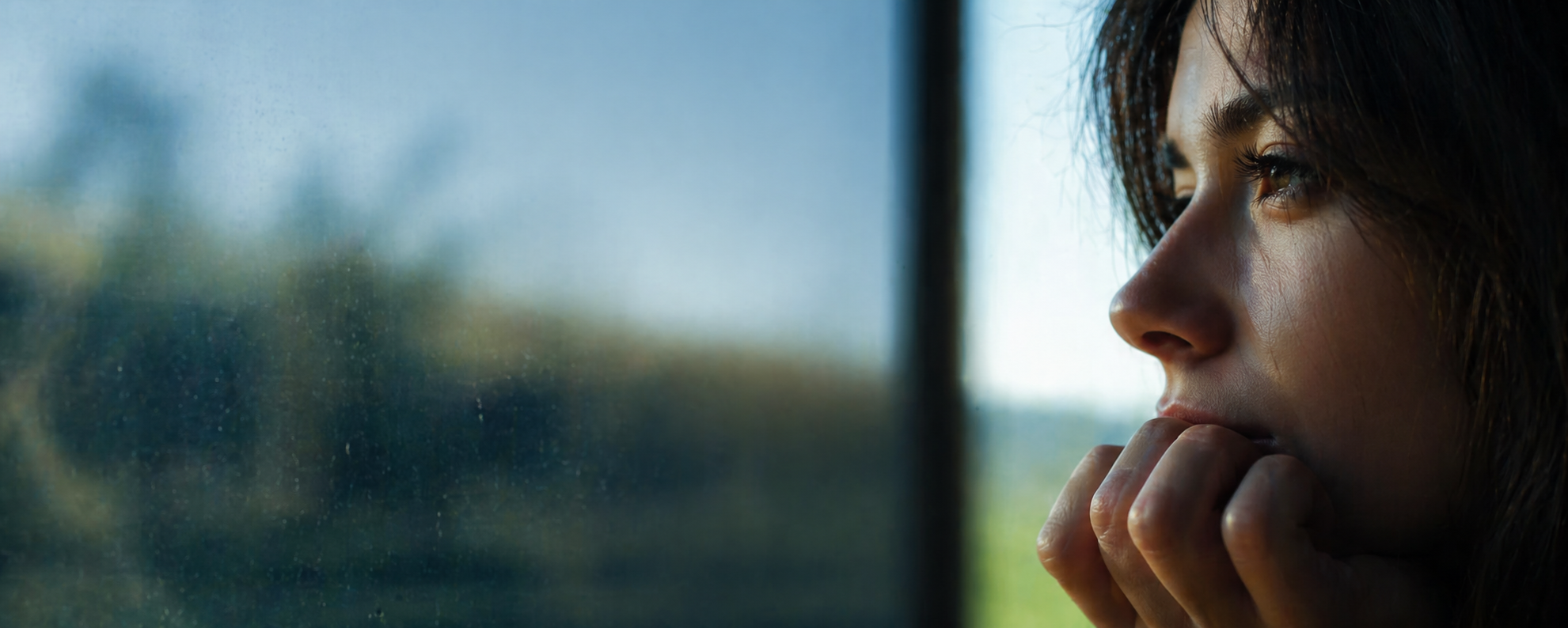 A woman with dark brown hair looking out a window with a contemplative expression, resting her chin on her hand.