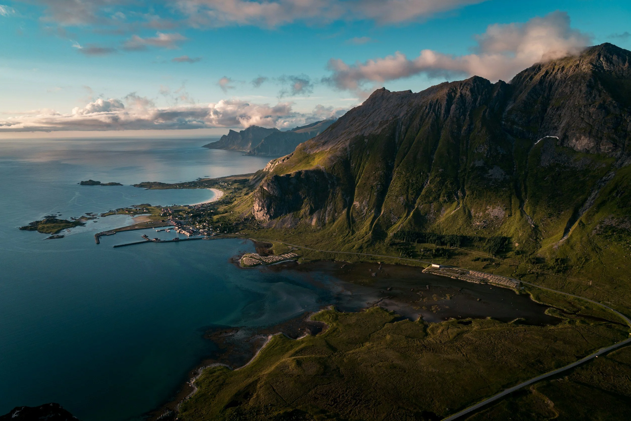 Aerial view of a rugged coastline with high green mountains, a bay, and a small town with circular harbor and boats.