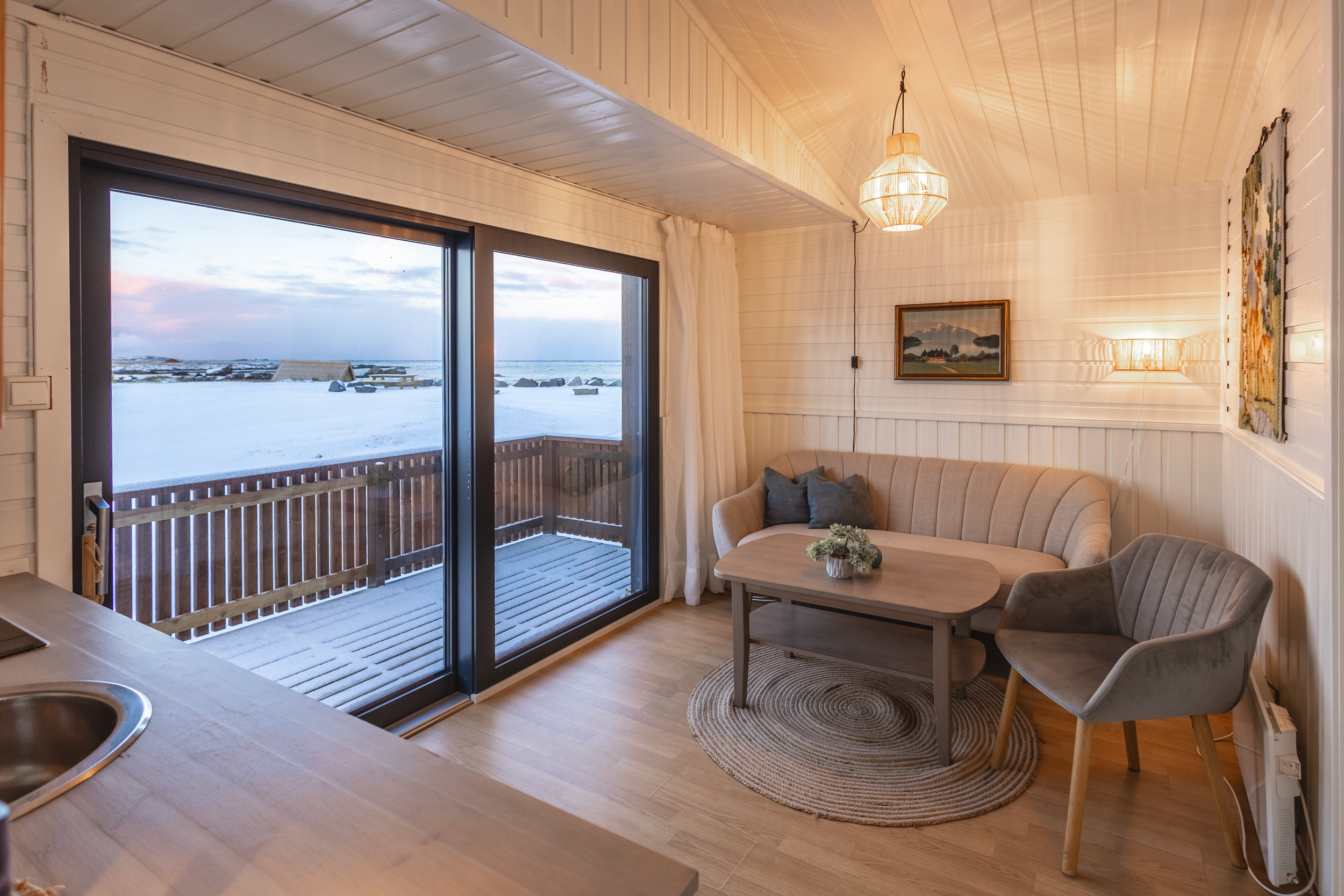 Cozy living room with a beige sofa, a grey armchair, a round wooden coffee table with three small decorative pumpkins, cream curtains, a woven ceiling light fixture, and ocean view through windows.