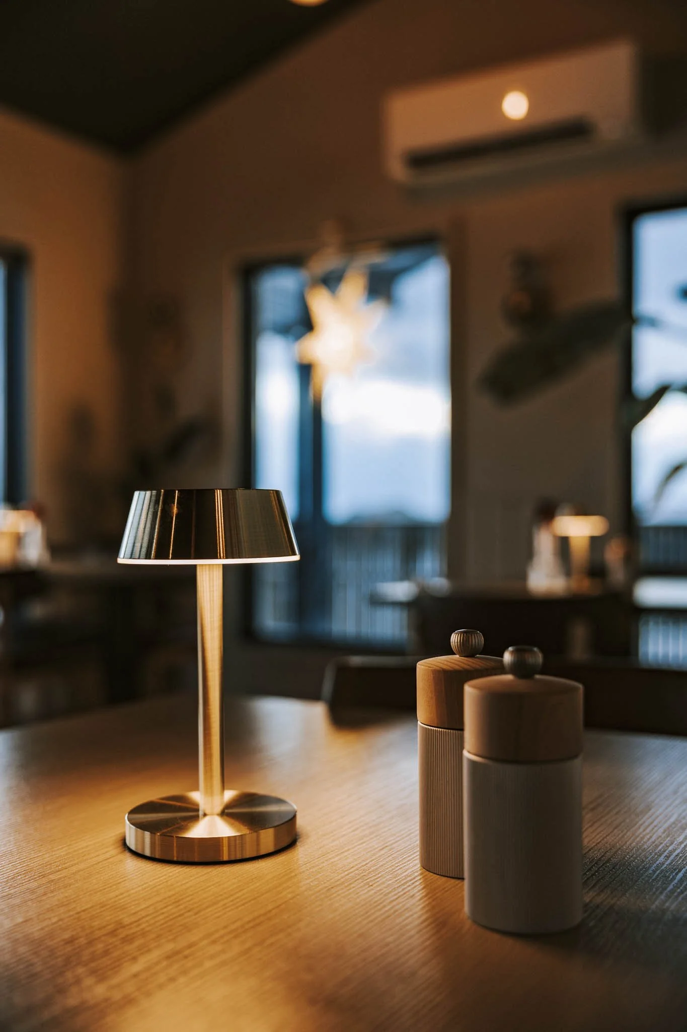 A table lamp and two wooden jars with knobs on a wooden table inside a cozy room with windows showing an outdoor view.