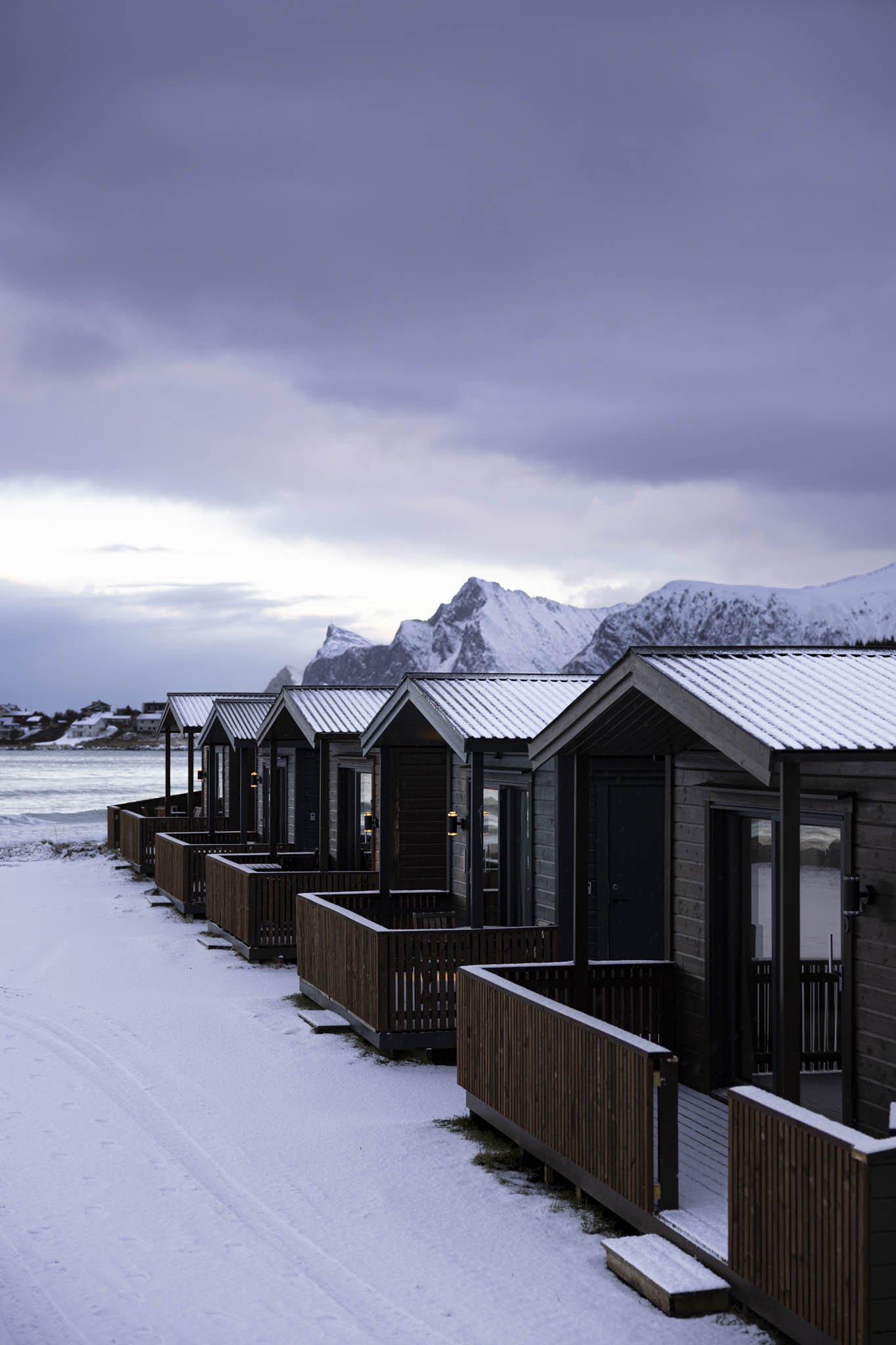 A row of dark wooden cabins with snow-covered roofs and small balconies, set against a snowy landscape with mountains and a cloudy sky in the background.