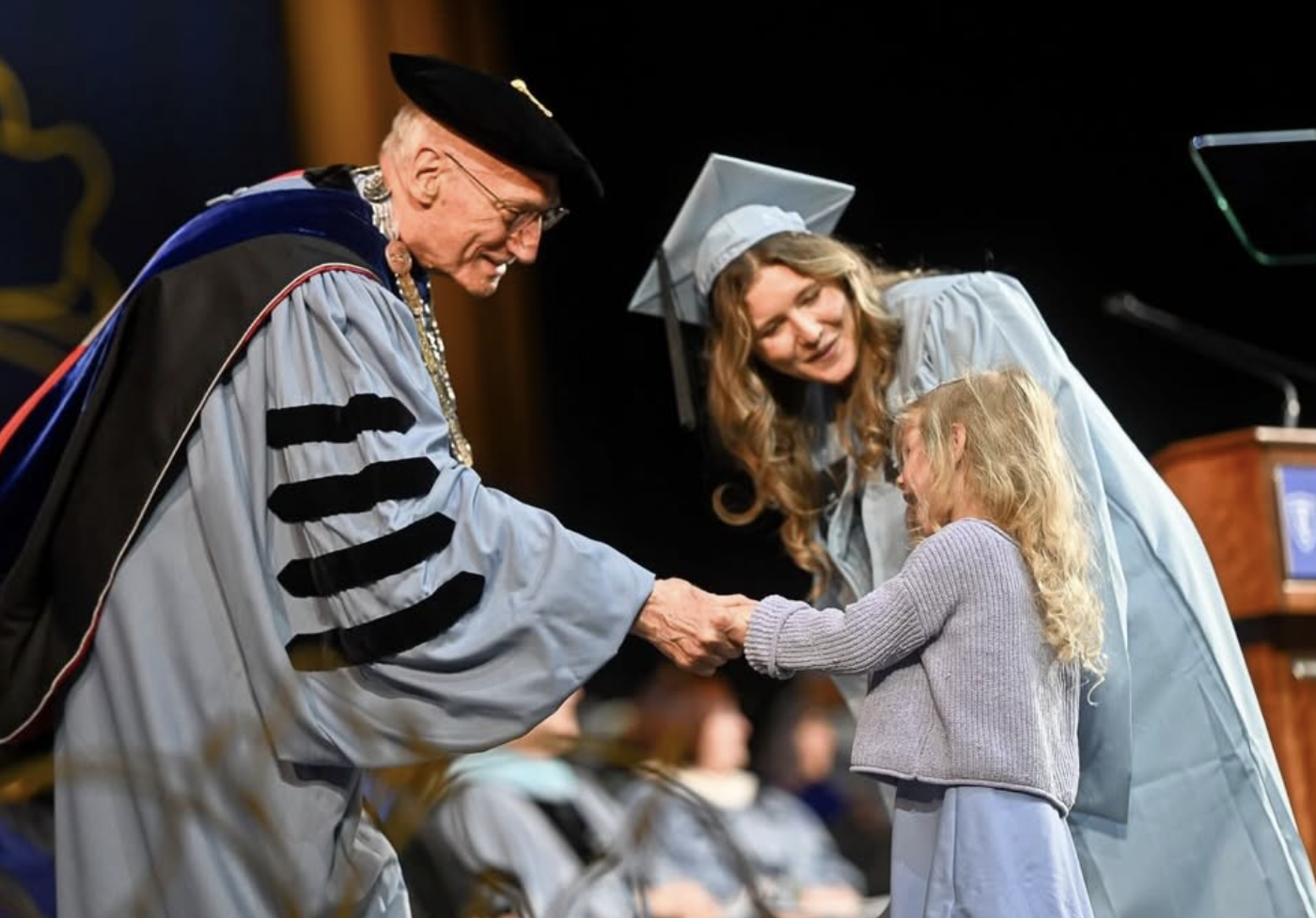 A graduation ceremony where an elderly man in academic regalia shakes hands with a young girl, while a woman in a graduation cap and gown looks on happily.