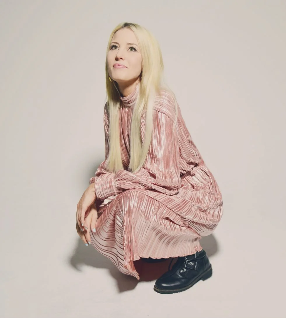 A woman with long blonde hair squatting against a plain wall, wearing a pink pleated dress and black shoes, looking upwards.