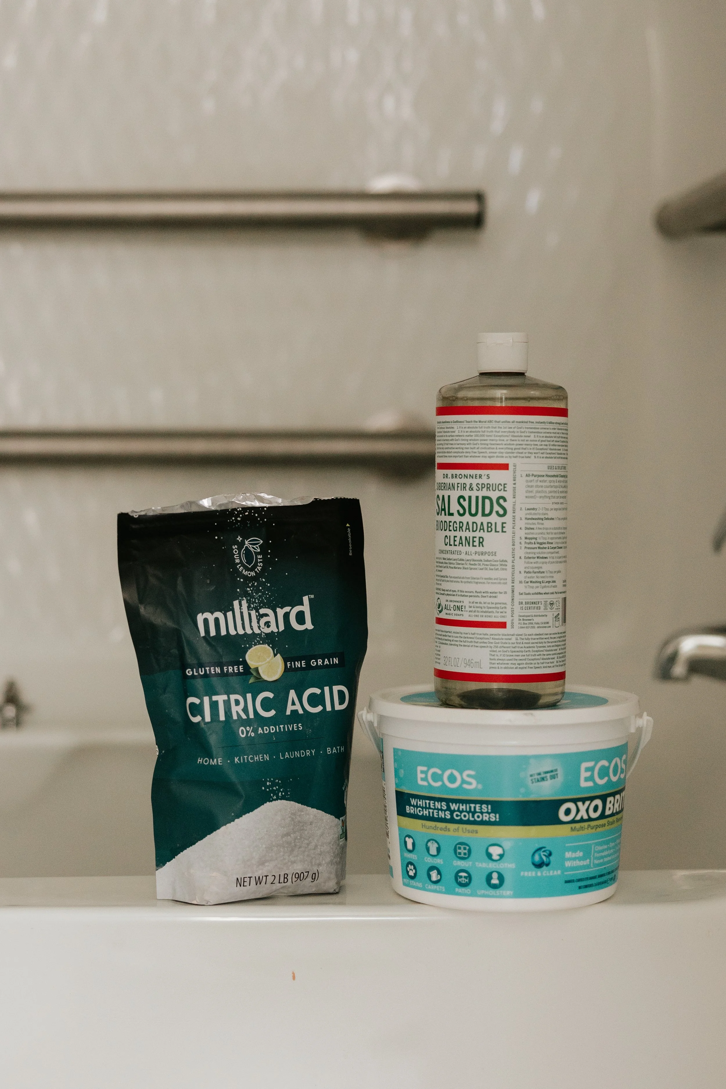 Cleaning supplies on a white surface with a washing machine in the background. The supplies include a bag of Mullard citric acid, a container of ECOS Oxi Bright, and a bottle of Dr. Bronner's Sal Suds biodegradable cleaner.