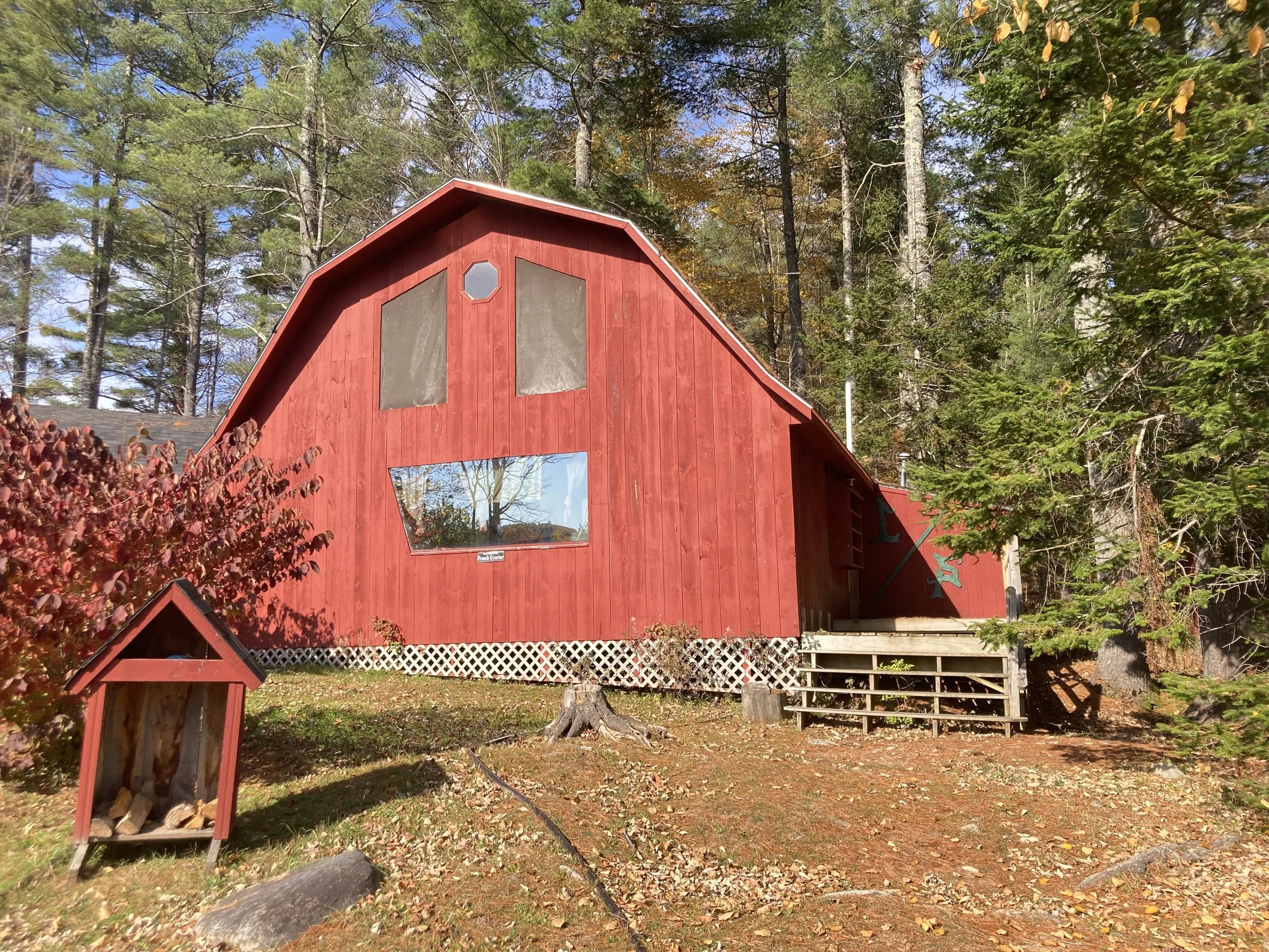 Red barn in a wooded area with stairs leading up to the entrance and a small shed in front, surrounded by trees and fallen leaves.