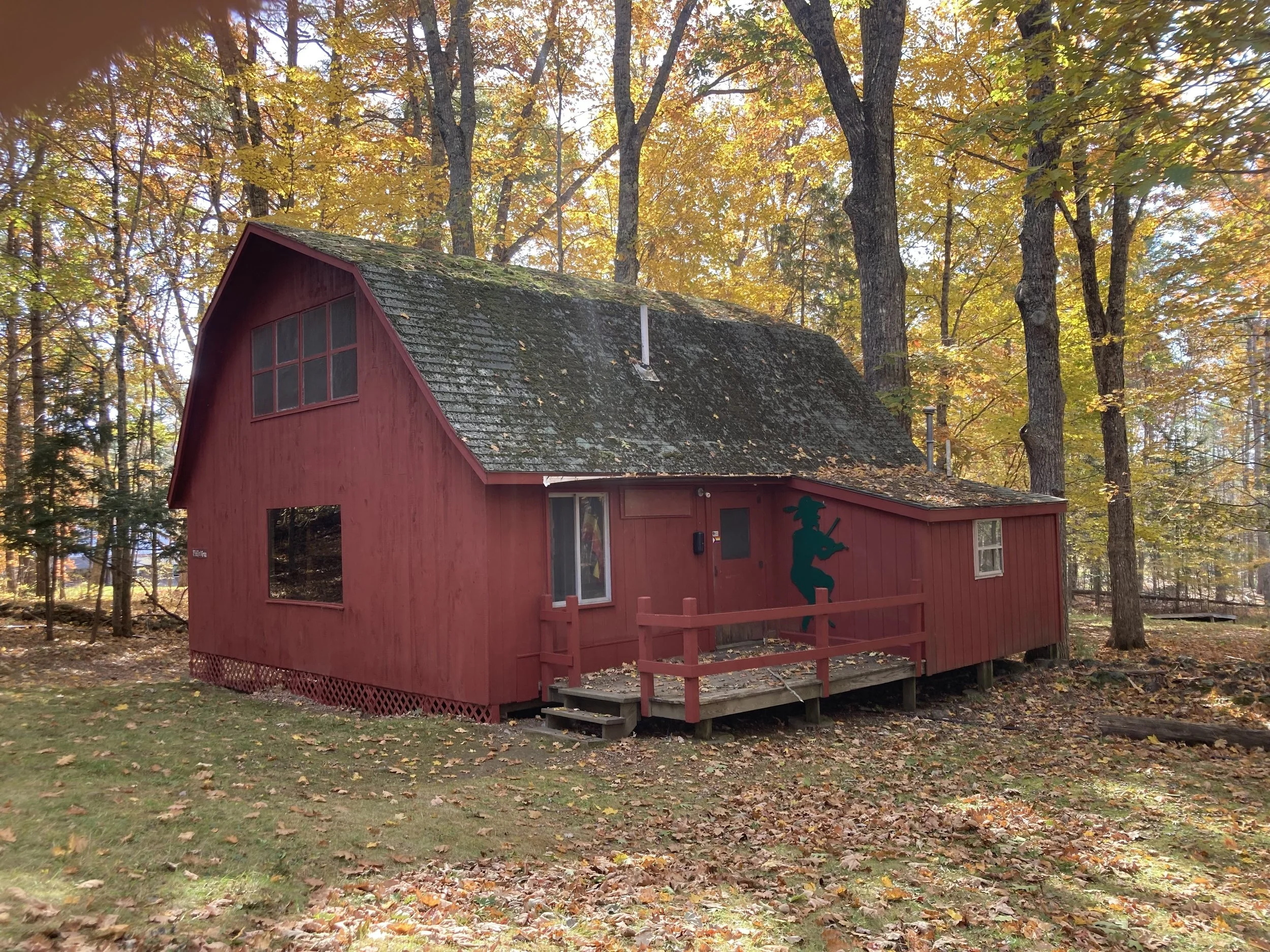 A red wooden house in the woods with a painted silhouette of a fiddler on the side. The house has a small porch, trees with fall foliage around, and moss on the roof.