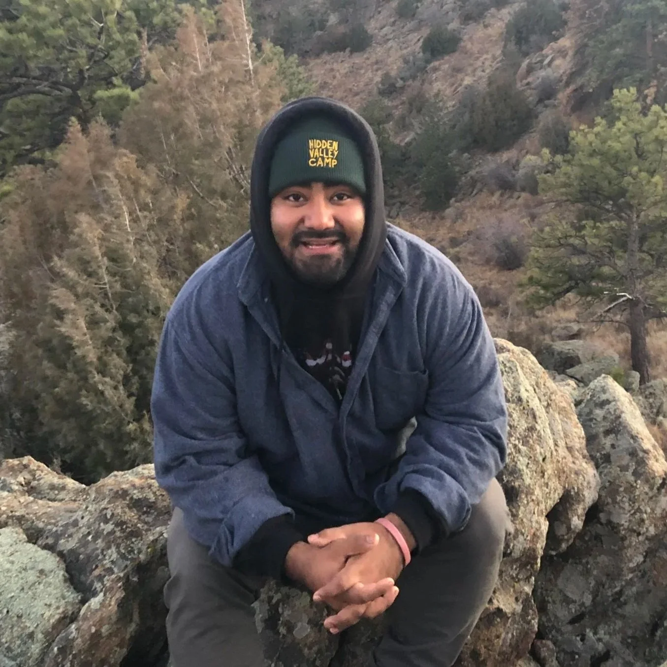 A man wearing a dark green beanie that says 'HIDDEN VALLEY CAMP' and a dark blue jacket, sitting on rocks outdoors with trees and hills in the background.