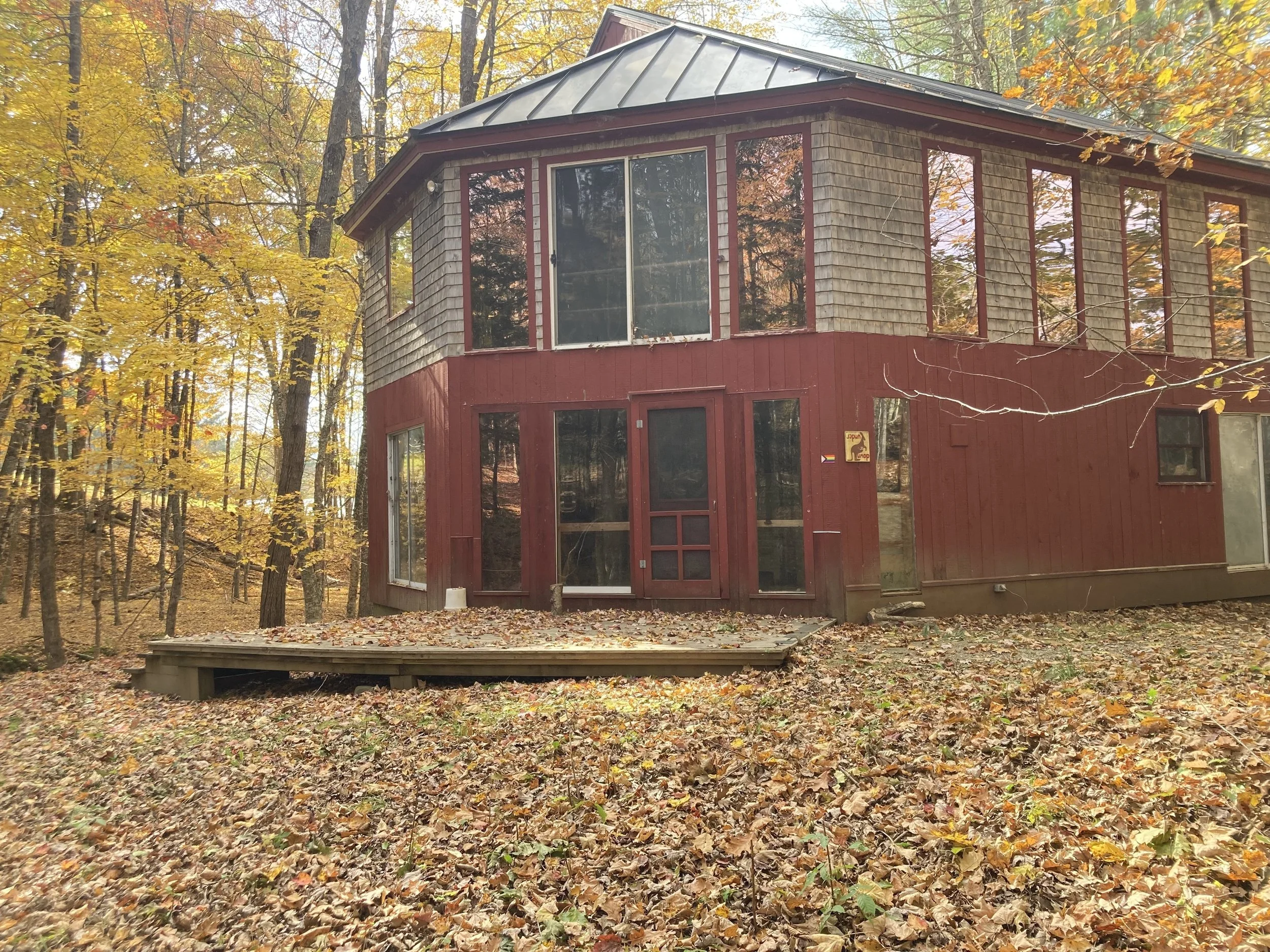 A two-story house with a red and tan wooden exterior, surrounded by fallen autumn leaves and yellow-orange trees.