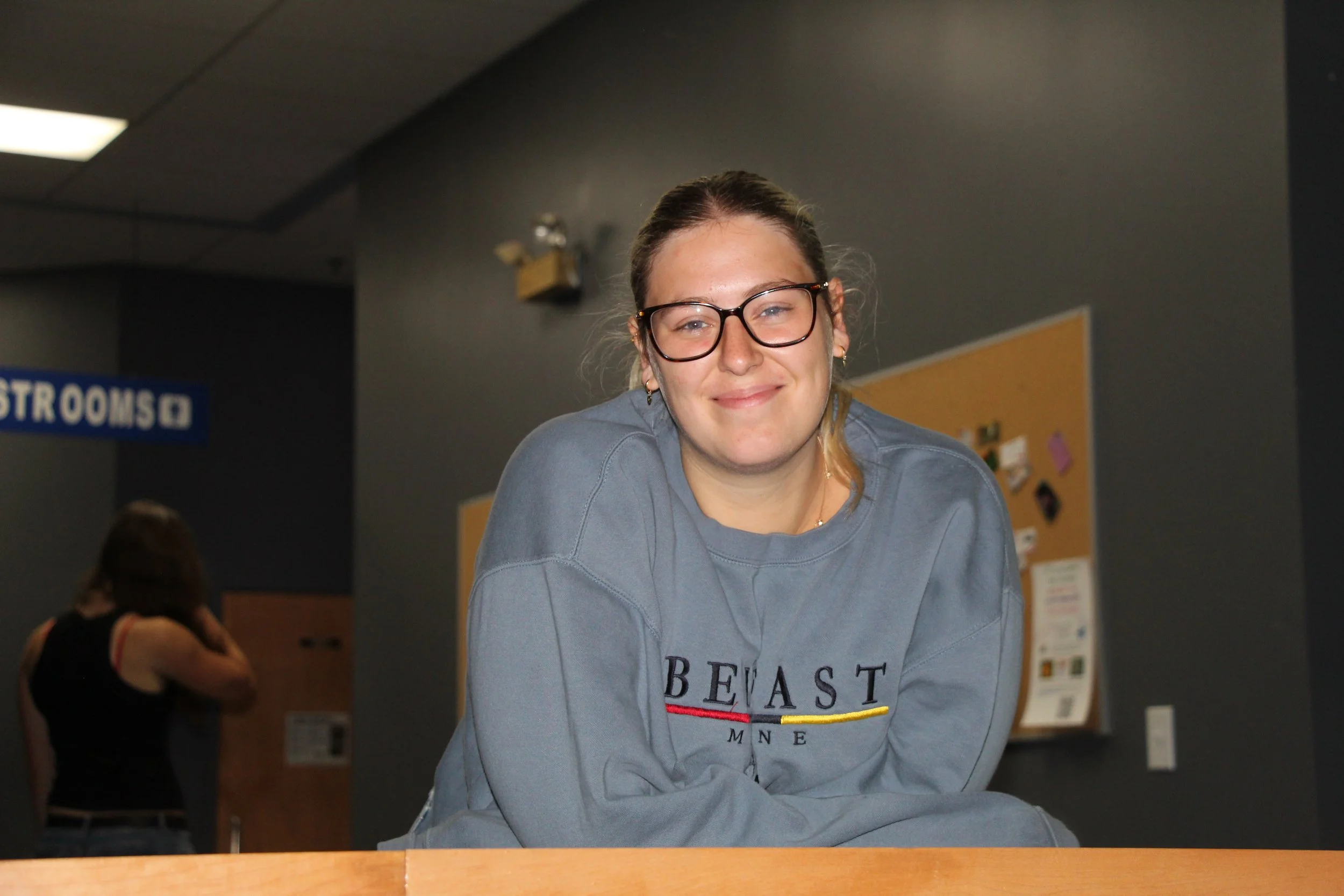 A woman with glasses and light brown hair tied back, smiling, wearing a gray sweatshirt with The Beast text, standing indoors against a gray wall, with a yellow bulletin board behind her. A sign indicating restrooms is visible in the background.
