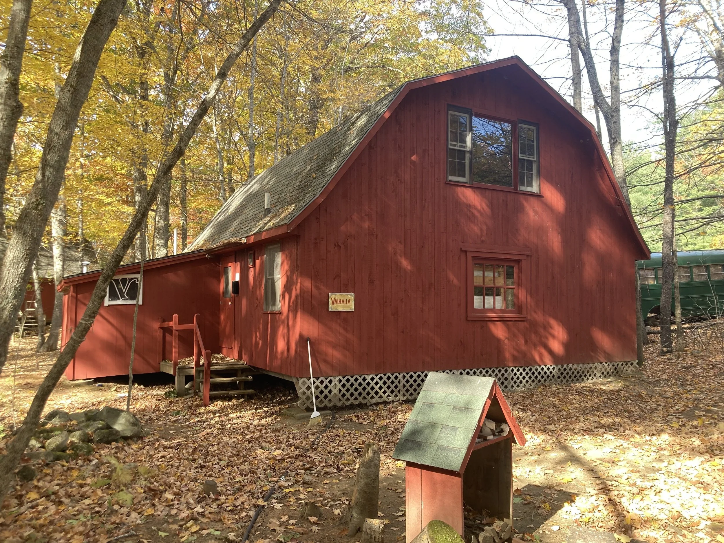 A red wooden cabin in a wooded area with fall foliage, featuring four windows and a sign that reads 'Vahalla' on the front, surrounded by leaves and a small wooden structure in the foreground.