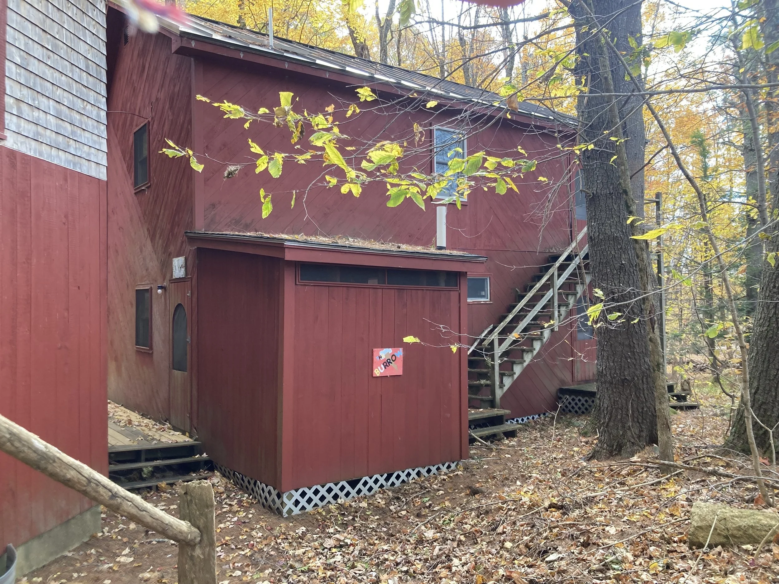 A red wooden house in a wooded area during fall. The house has exterior stairs leading to a second-floor entrance and small windows. Fallen leaves cover the ground, and trees with yellow leaves surround the house.