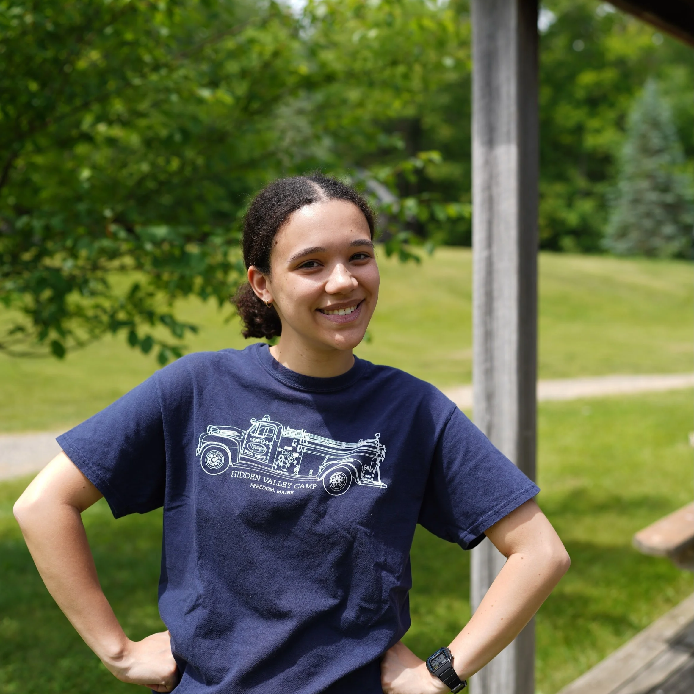 A smiling young woman with curly hair wearing a navy T-shirt with a graphic of a vintage truck, standing outdoors in a park with green trees and grass in the background.